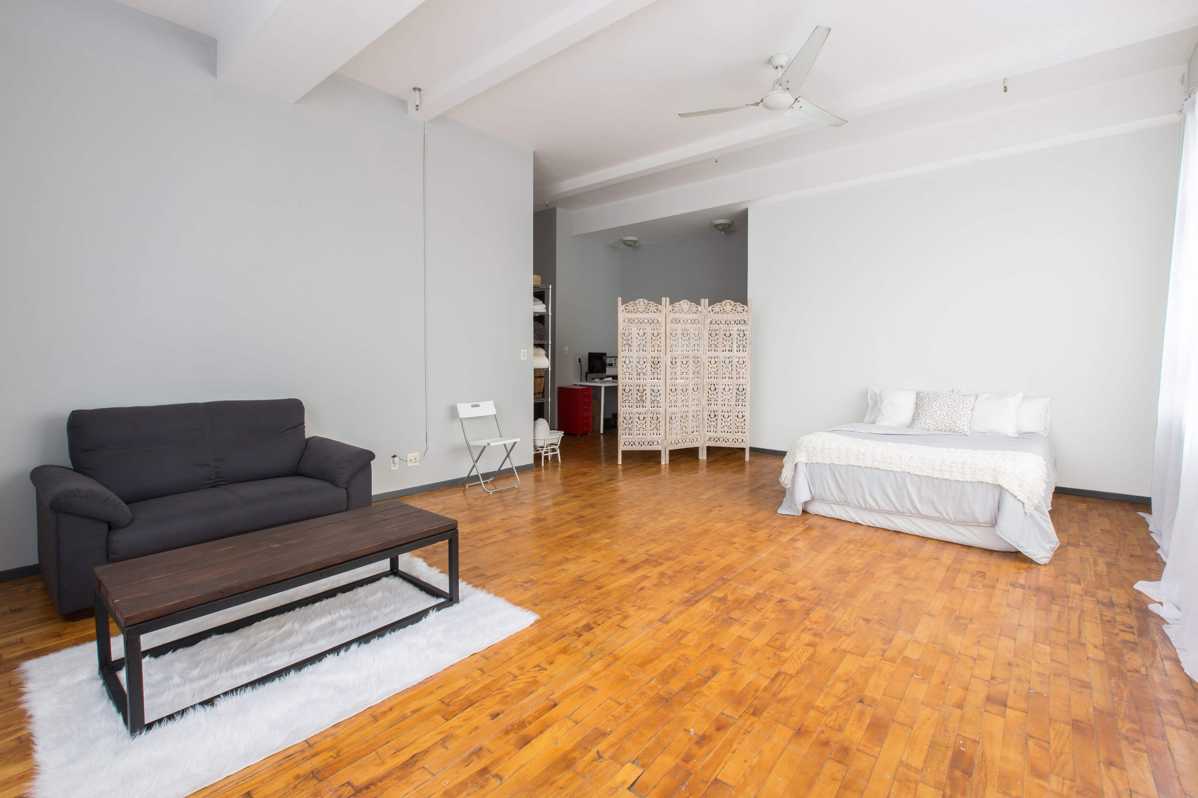 The image shows a spacious, minimally furnished living area featuring a black couch, a wooden coffee table on a white rug, a folding chair, a screen divider, and a bed in a bright room with wooden flooring.