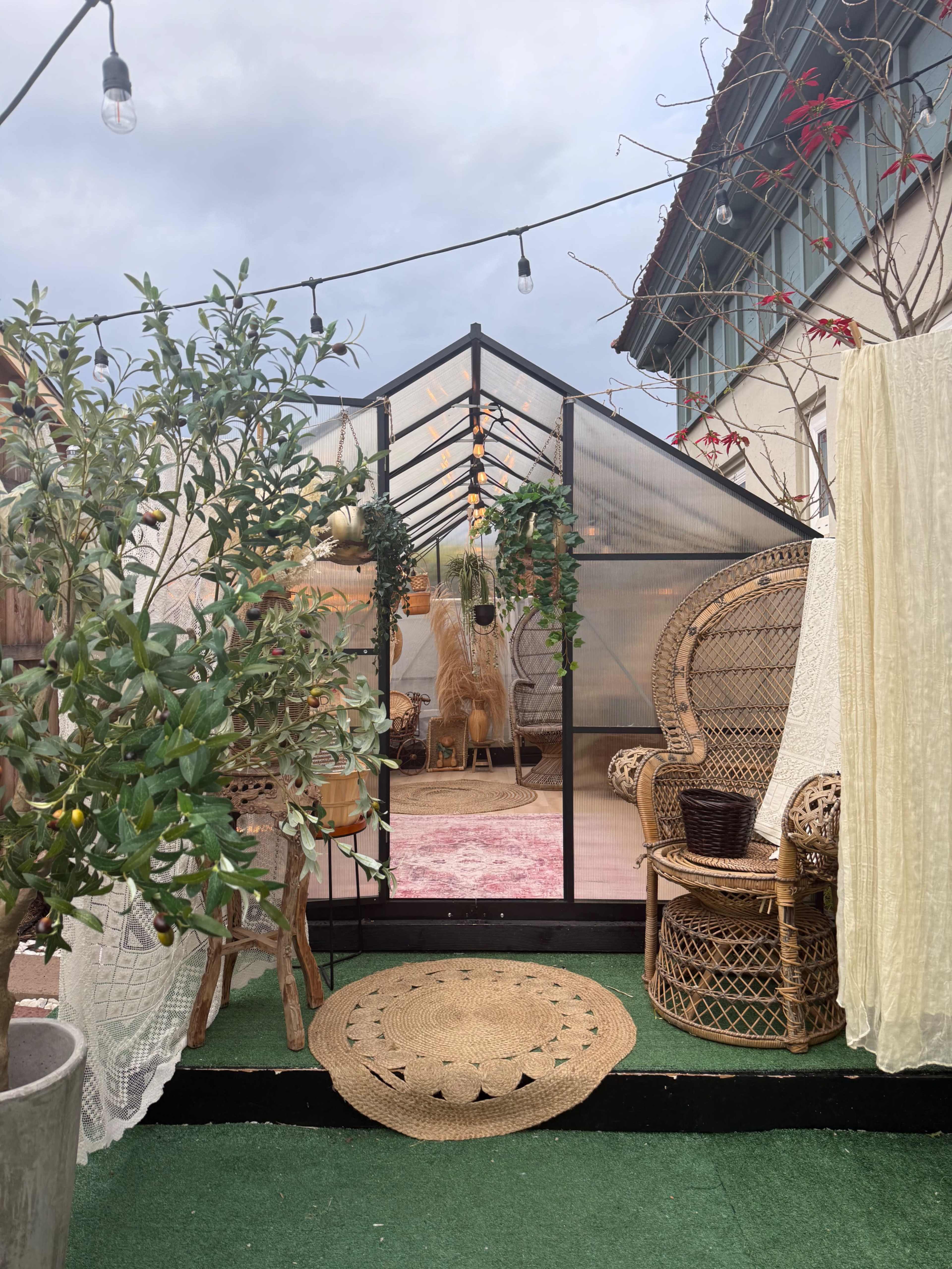 The image shows a greenhouse with a cozy interior featuring decorative plants, a wicker chair, and string lights overhead.