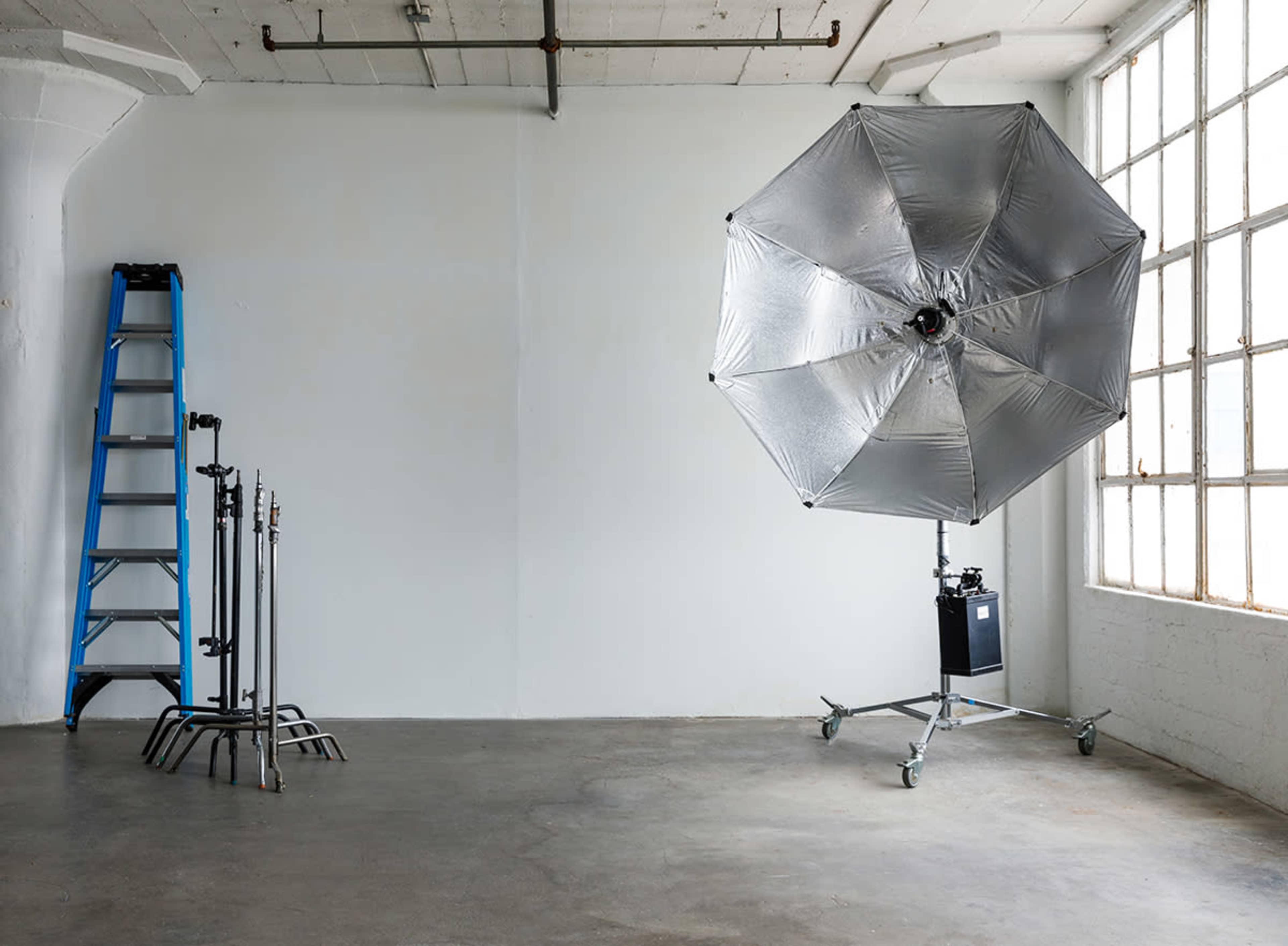 The image shows a spacious, empty studio with a large silver umbrella light on a stand and a blue ladder leaning against the wall.