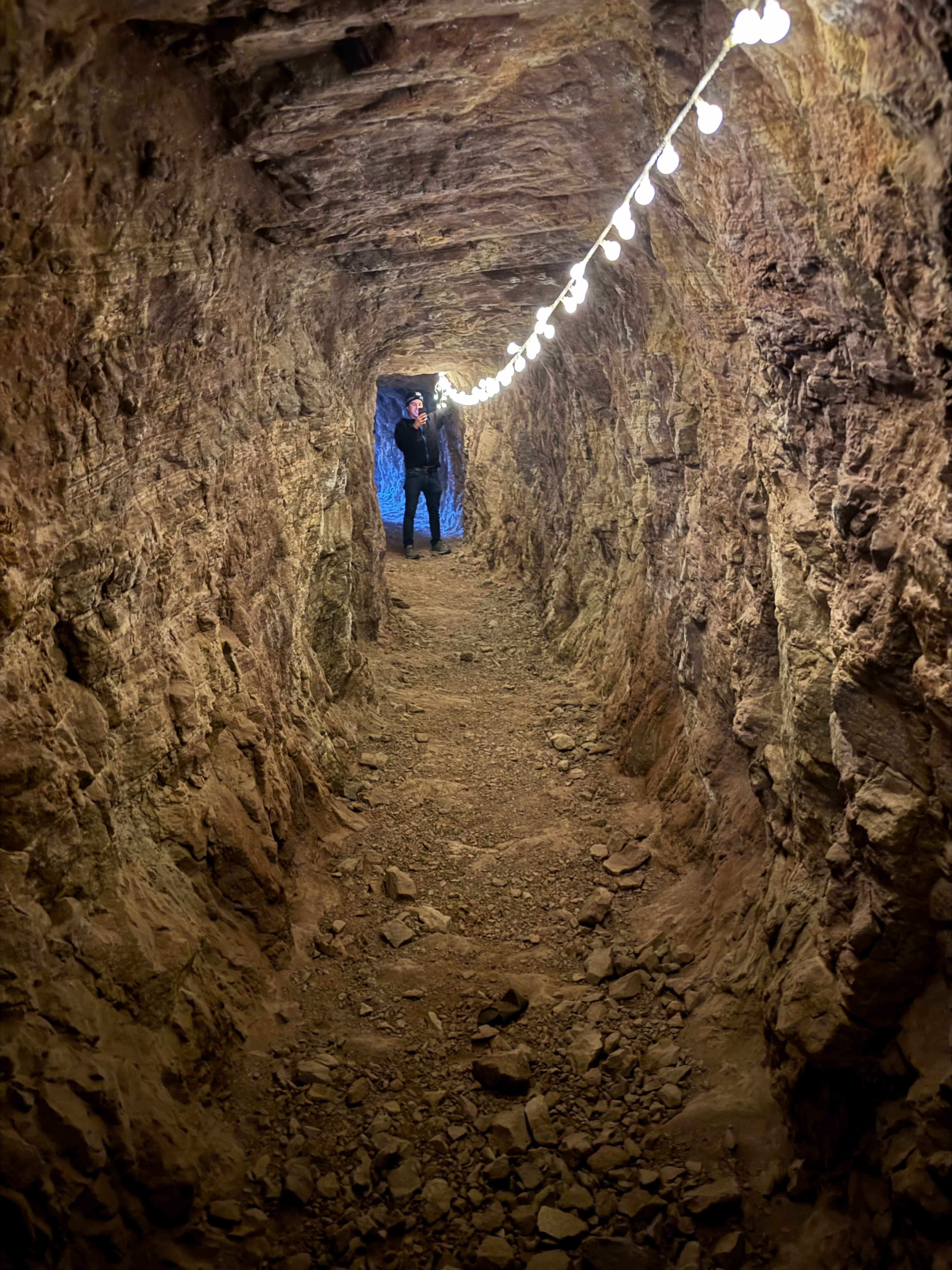 A narrow tunnel with rough stone walls is illuminated by a string of lights, leading toward a figure standing at the end.
