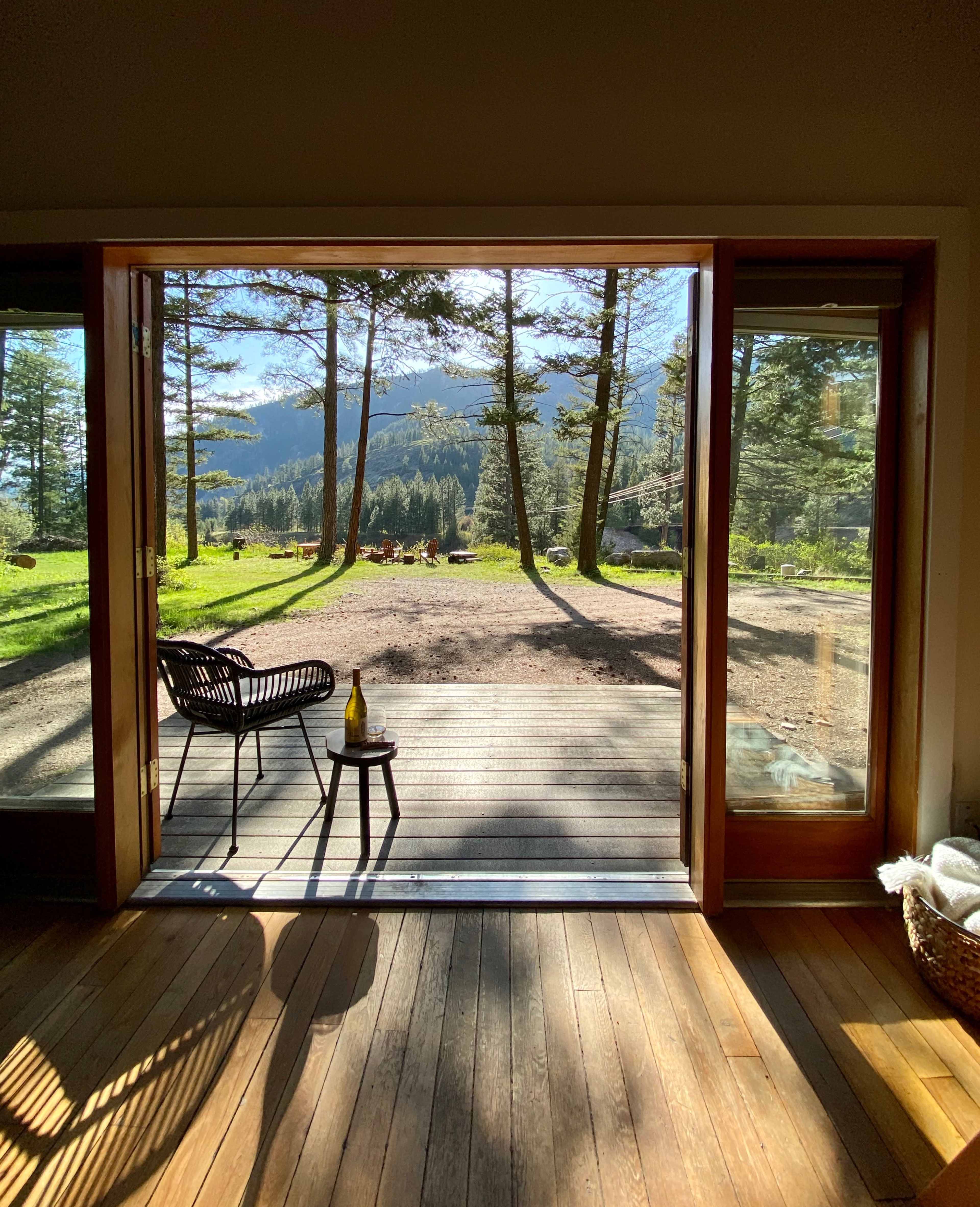 A wooden deck extends from an open doorway, framed by trees and mountains in the background.