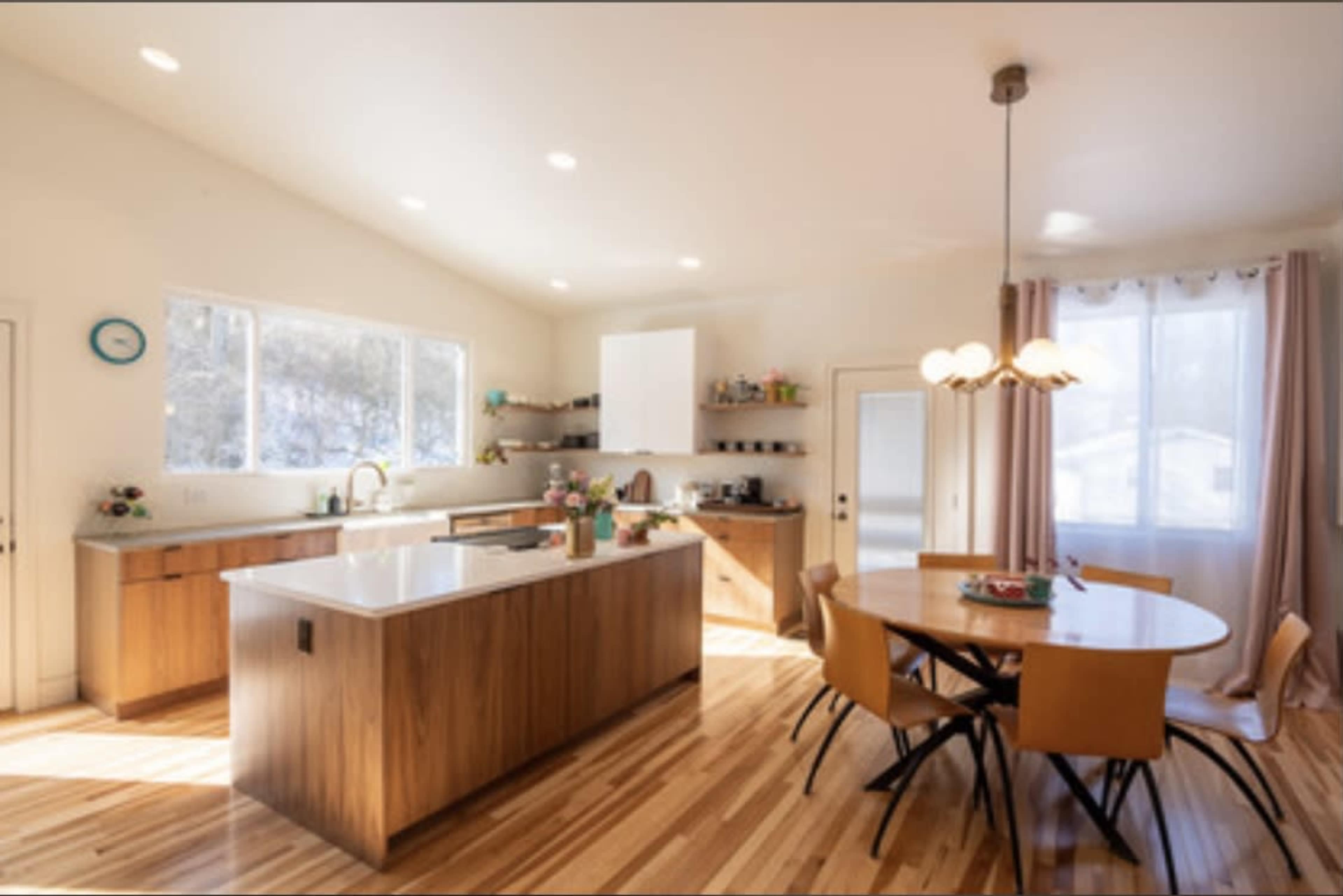 A modern kitchen features an island with a sink, wooden cabinetry, and a dining table surrounded by chairs, illuminated by natural light from large windows.