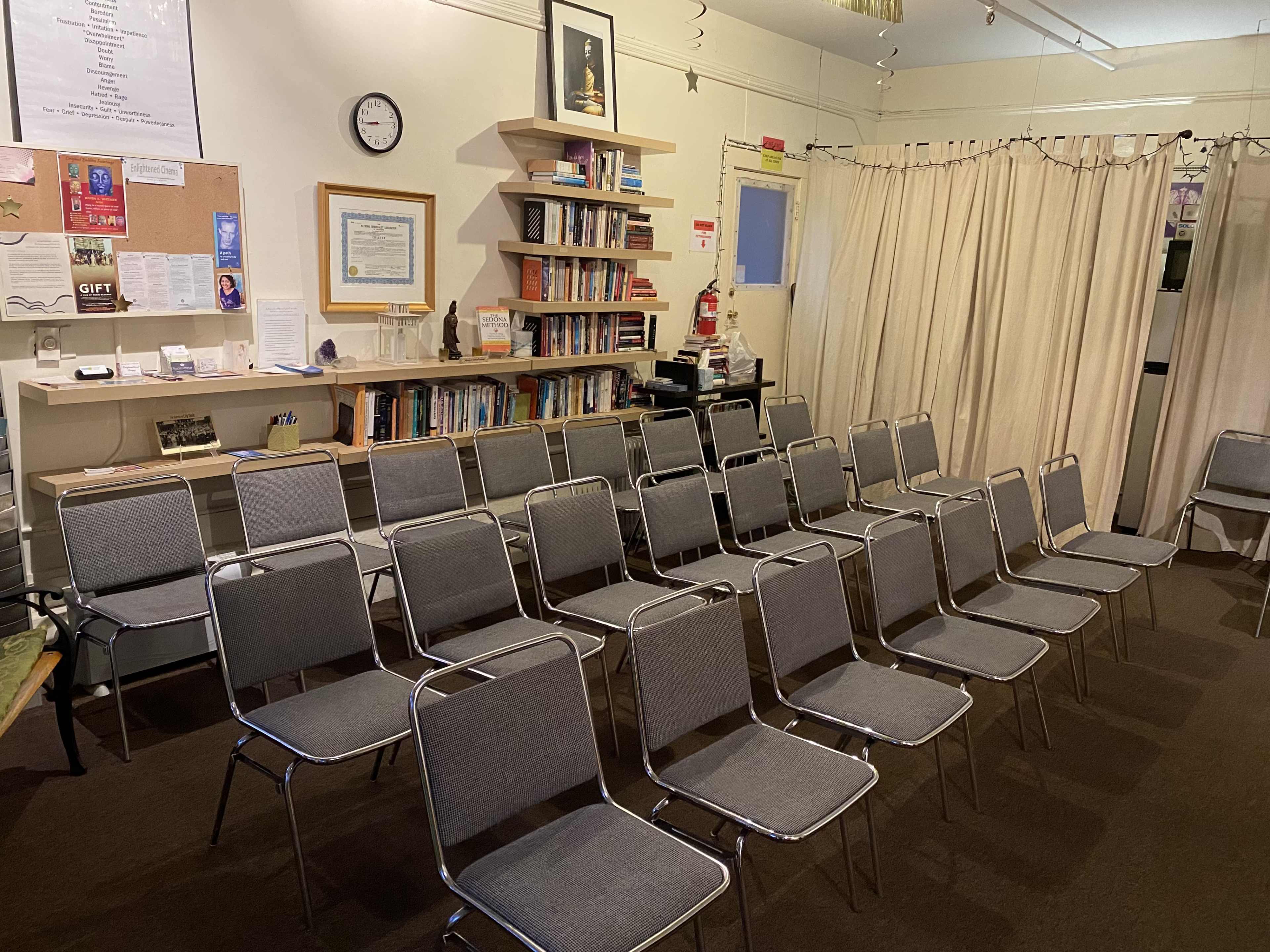 The image shows a room with several rows of grey chairs arranged facing a bookshelf and a bulletin board on one side.