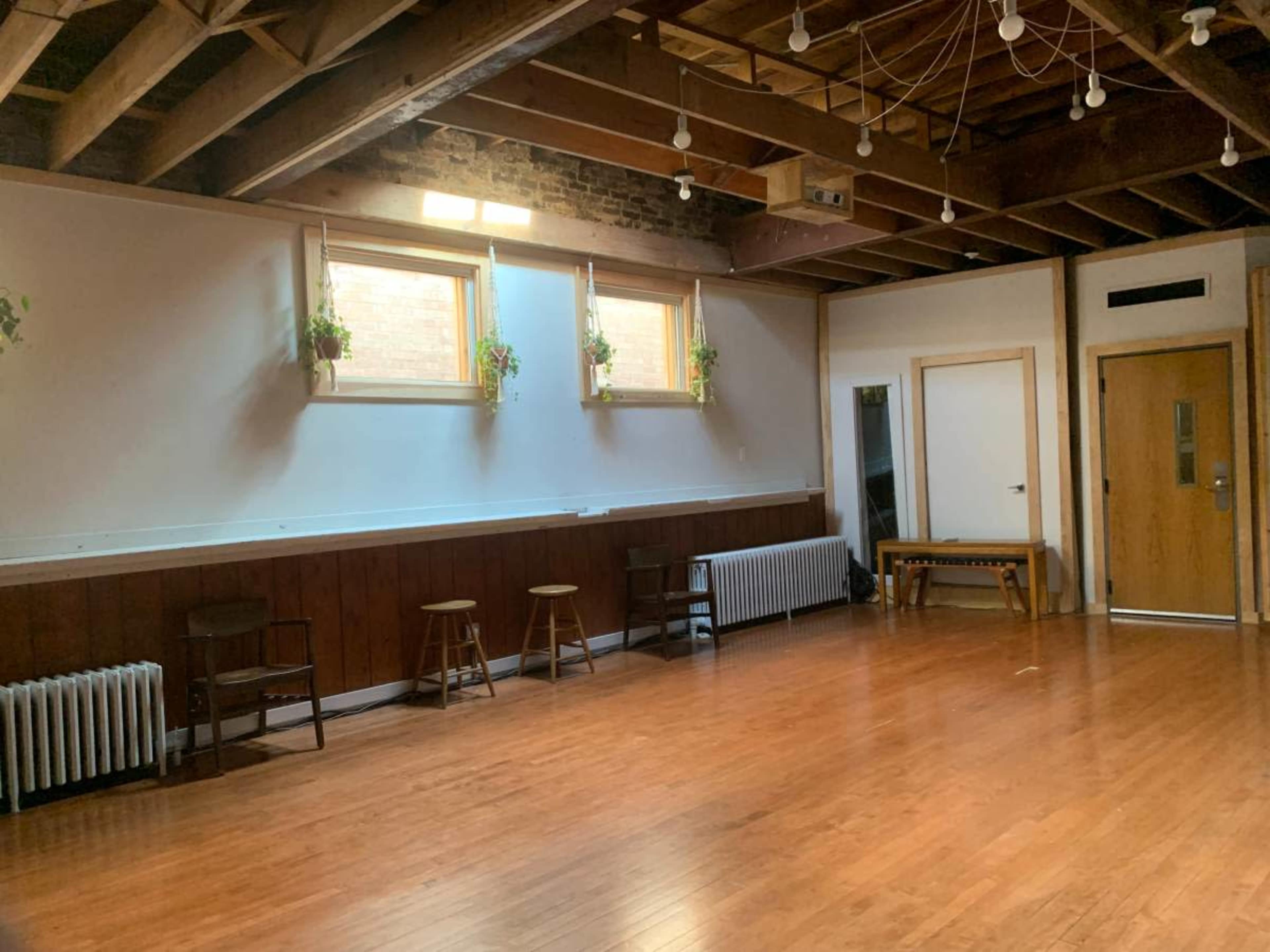 The image shows a spacious, empty room with wooden floors, exposed beams, and large windows featuring potted plants.