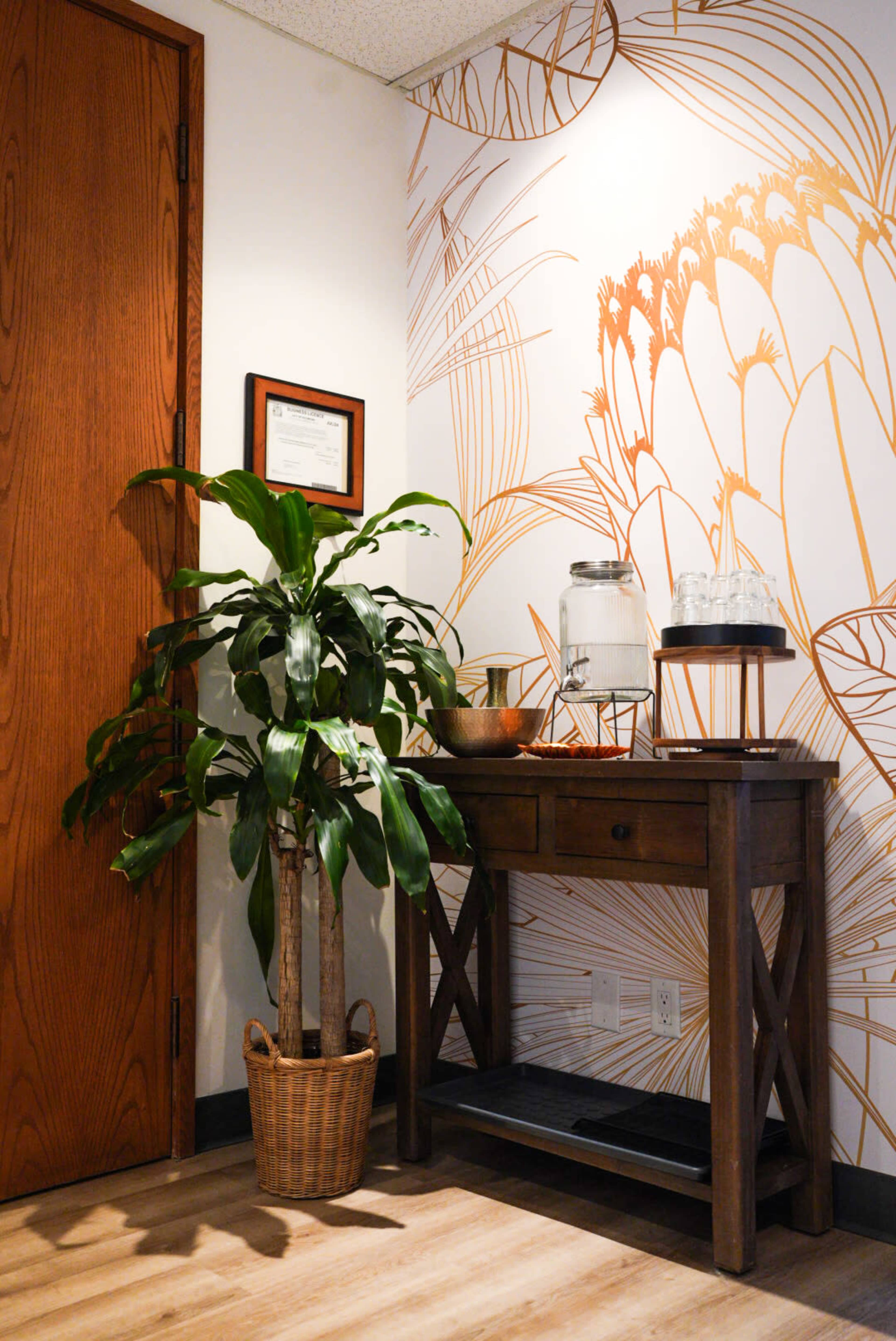 The image shows a corner of a room featuring a wooden console table beside a potted plant and a wall with a decorative floral mural.