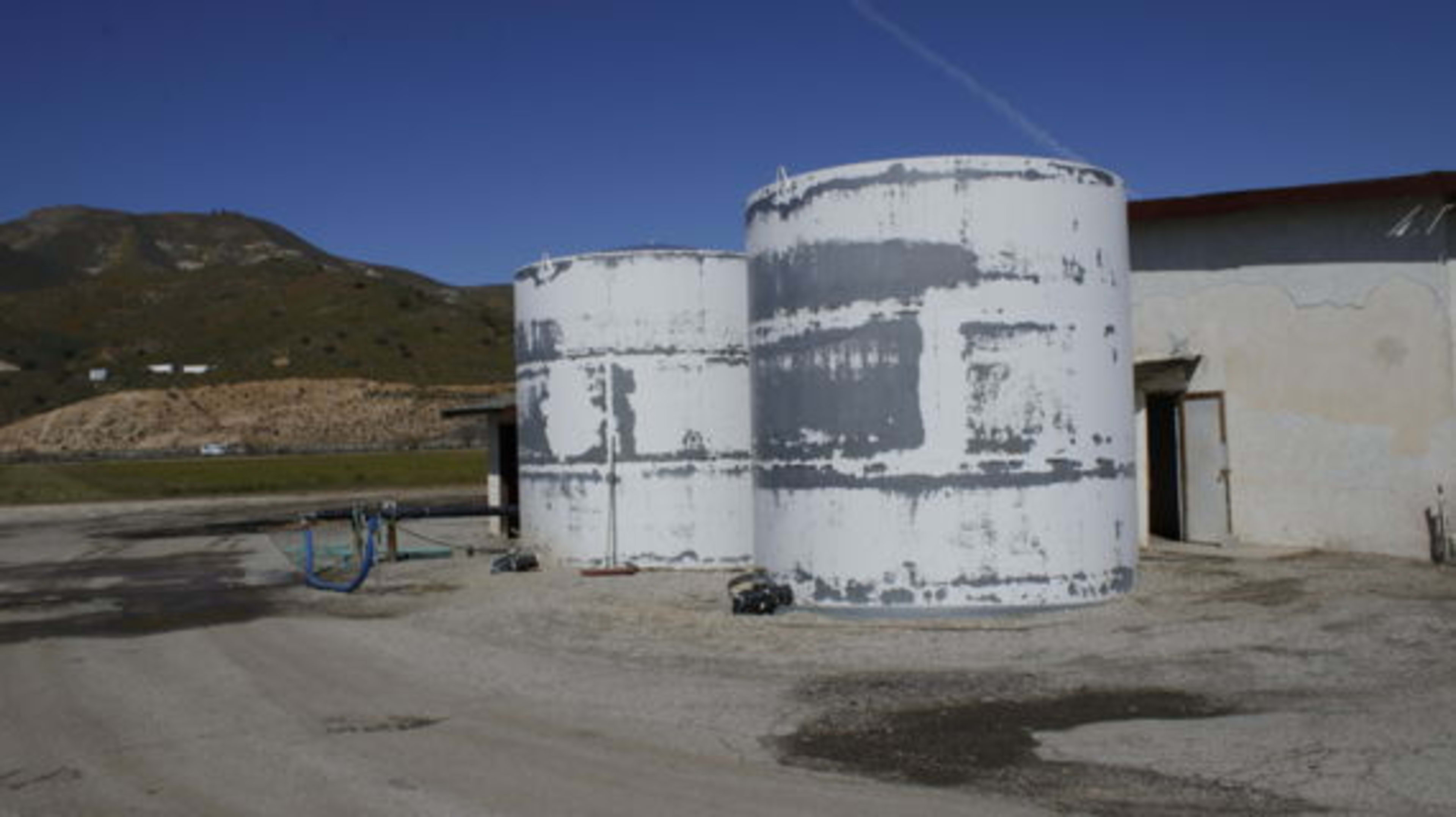 Two large, cylindrical storage tanks beside a building on a gravel surface, with a mountainous landscape in the background.
