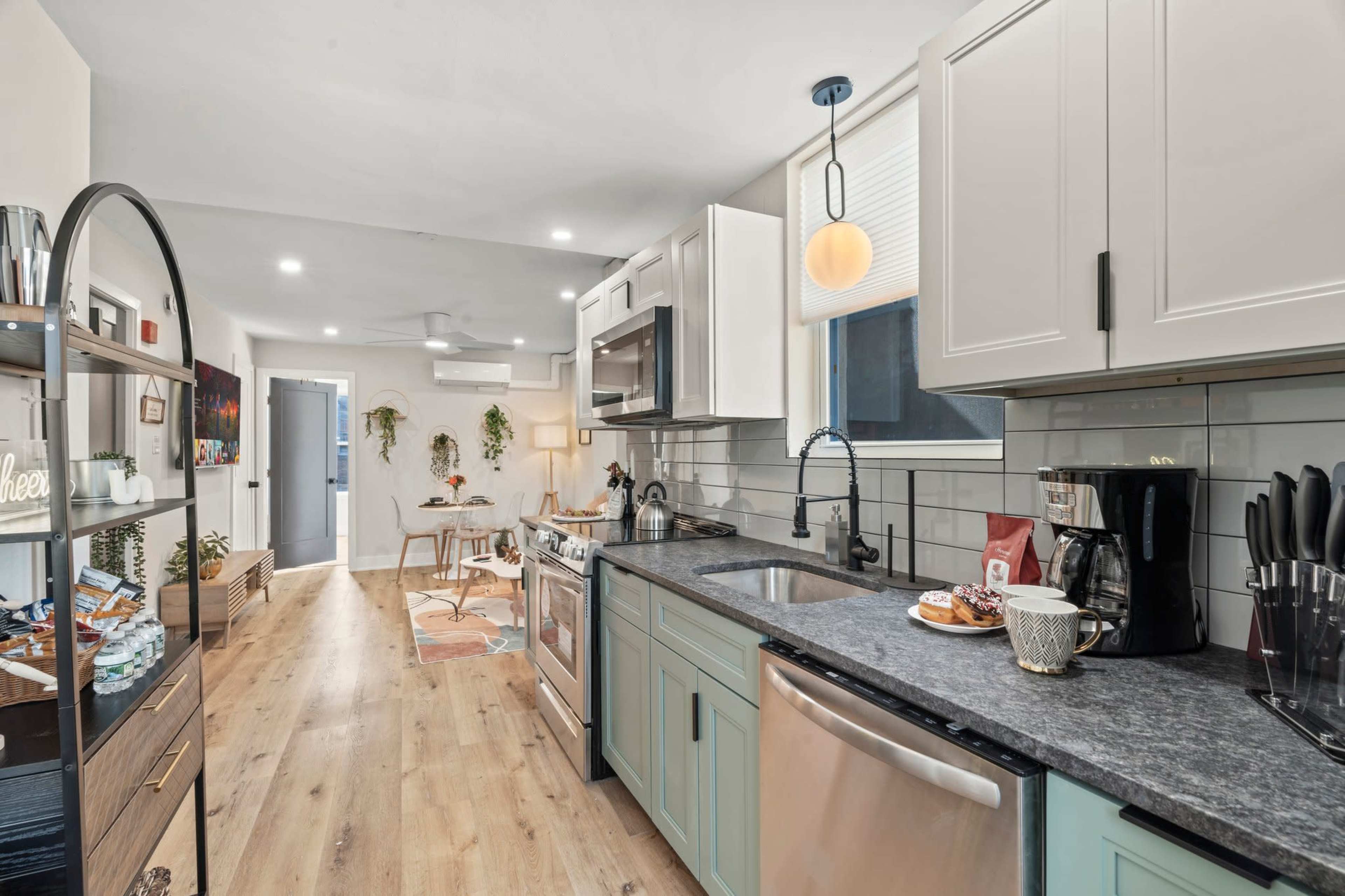 The image shows a modern kitchen with light cabinetry, a dark countertop, and stainless steel appliances, leading to an adjacent dining area.