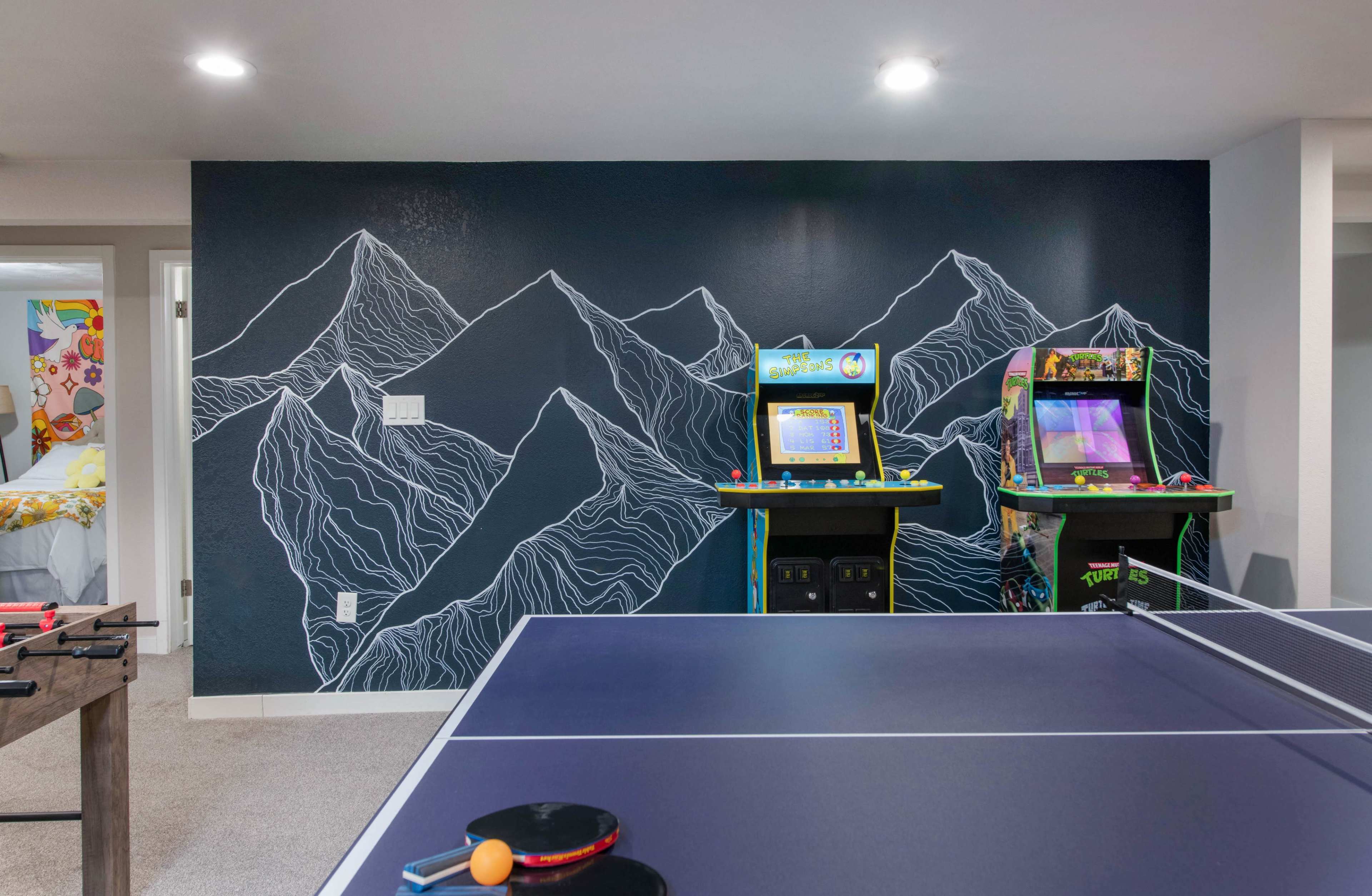 The image features a game room with a ping pong table in the foreground and two vintage arcade machines against a mountain-themed mural on the wall.