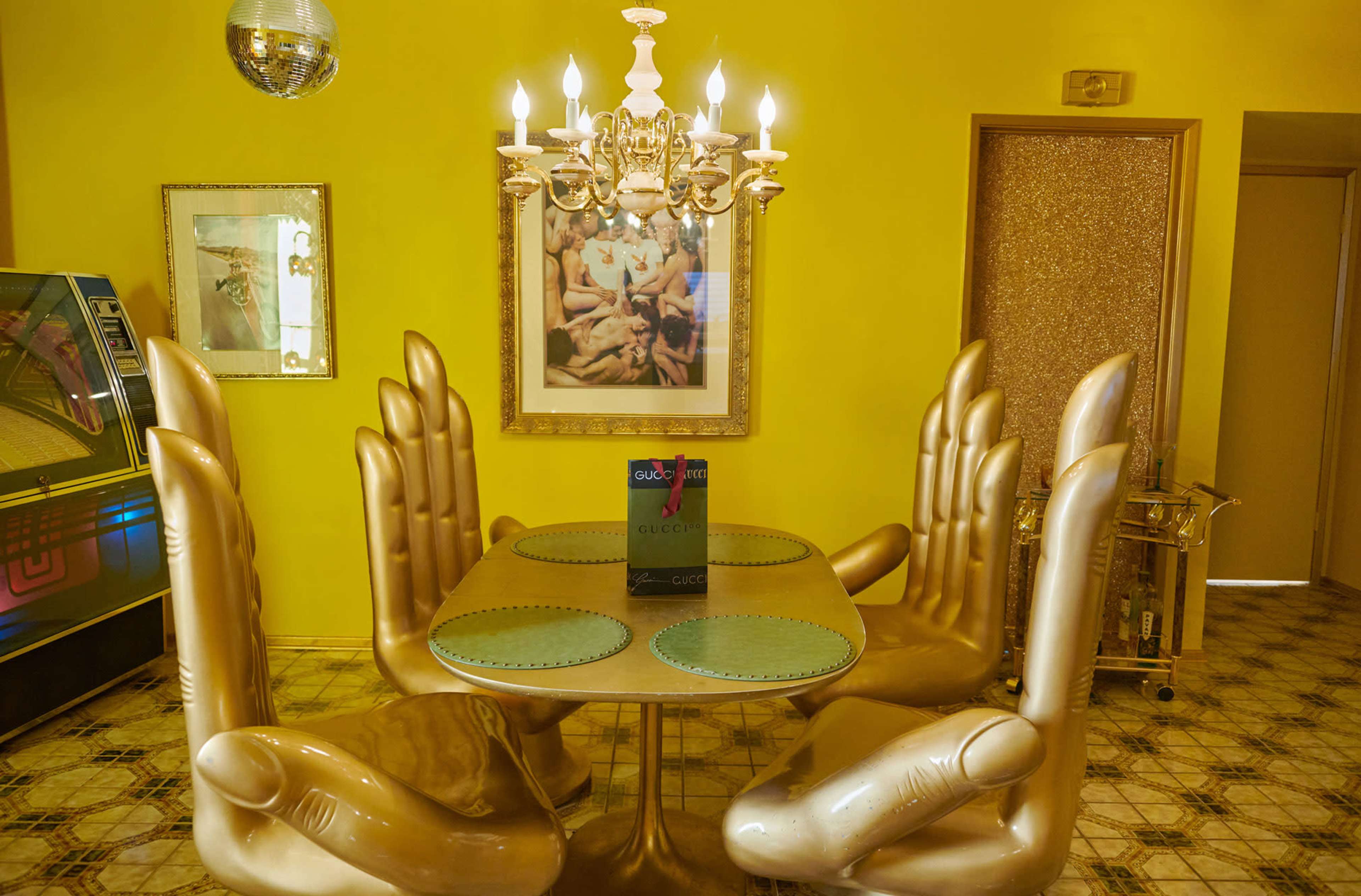 A round table with gold hand-shaped chairs is set in a brightly colored yellow dining room, featuring a chandelier and a framed artwork on the wall.