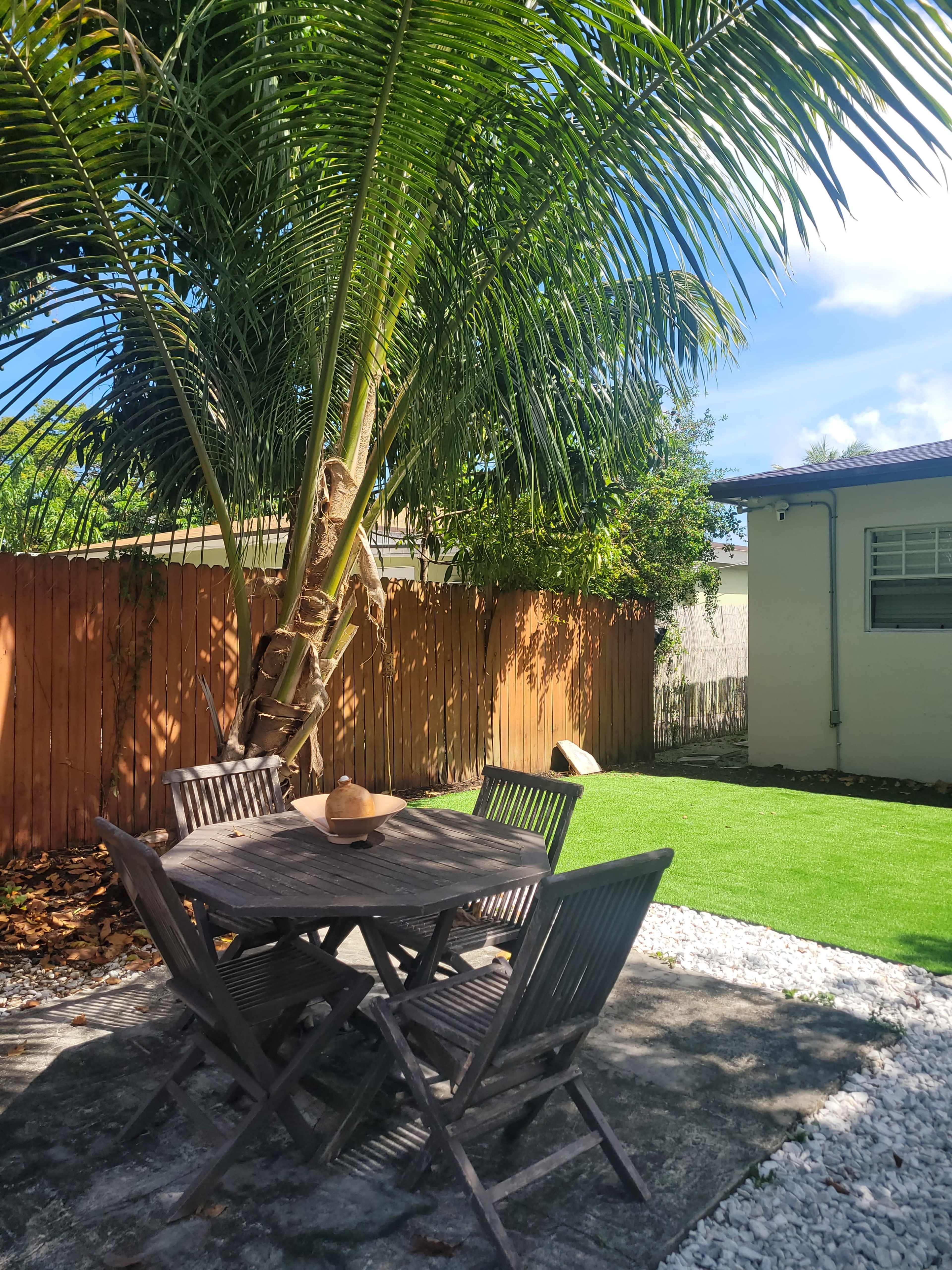 A wooden table with chairs sits on a stone patio under a palm tree in a backyard with a grassy area and a wooden fence.