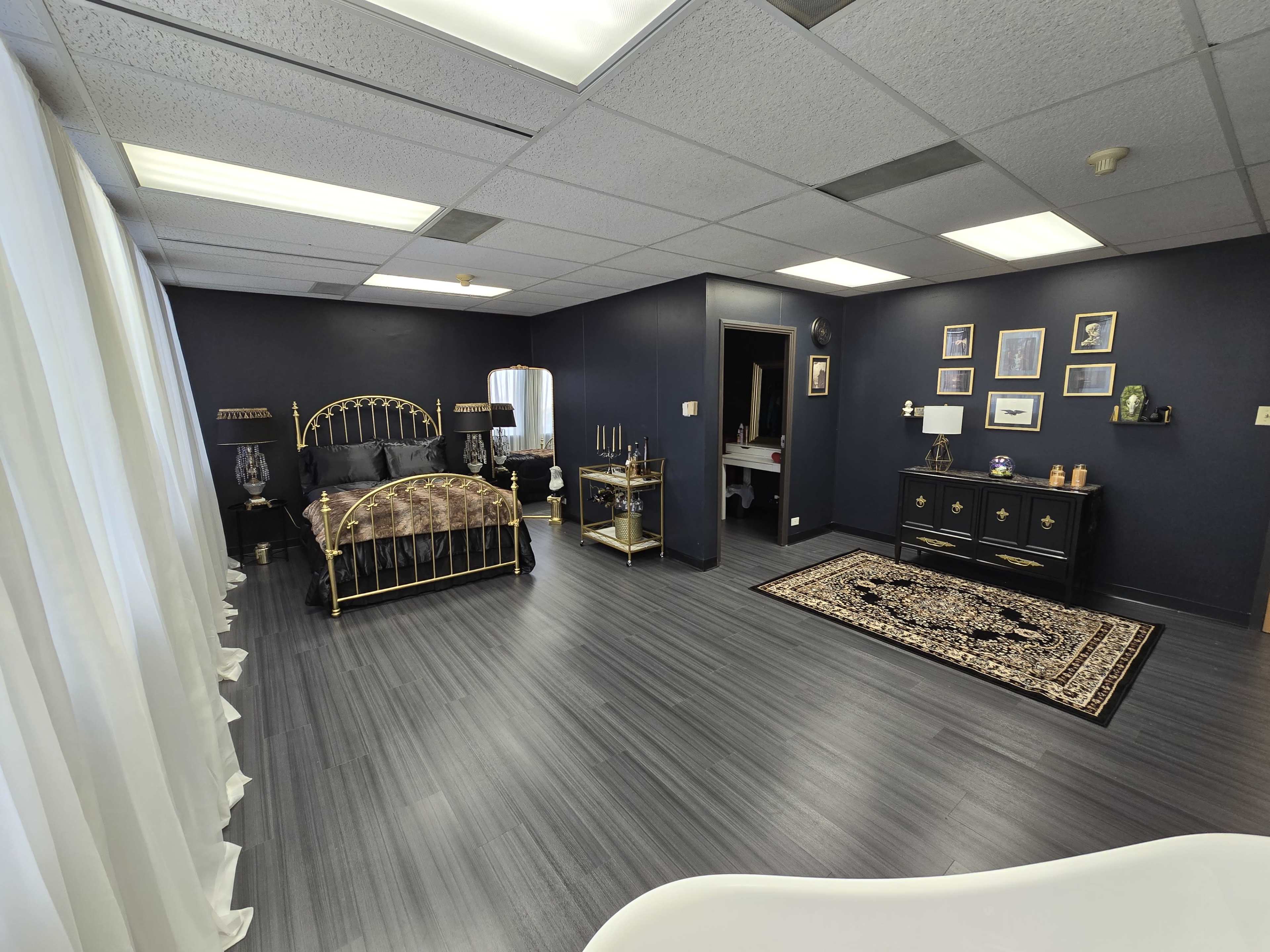A spacious bedroom featuring a gold bed frame, dark blue walls, a black dresser, and a decorative area rug.