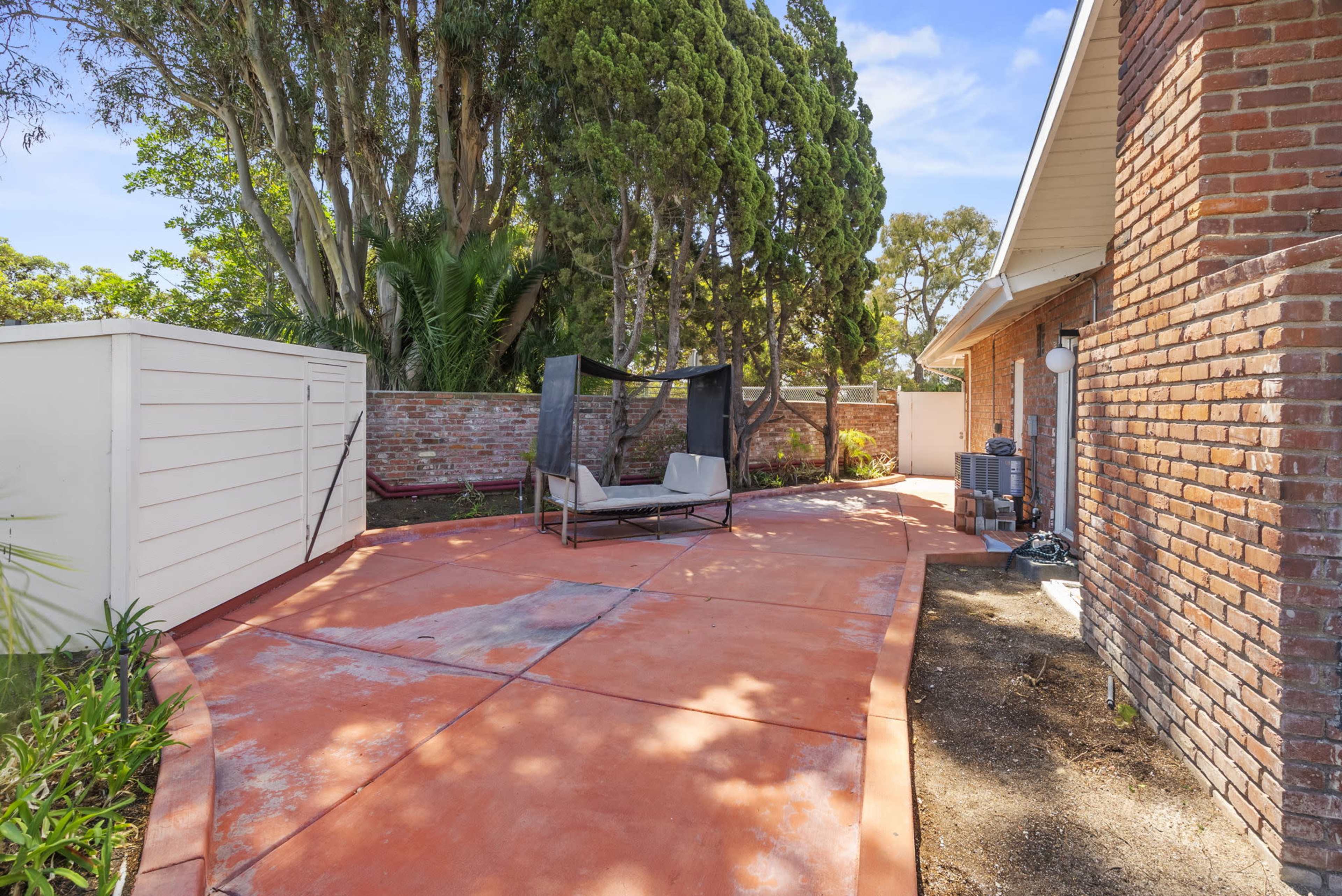 The image shows a patio area with a red concrete surface, featuring a shaded seating arrangement and surrounded by greenery and a brick wall.