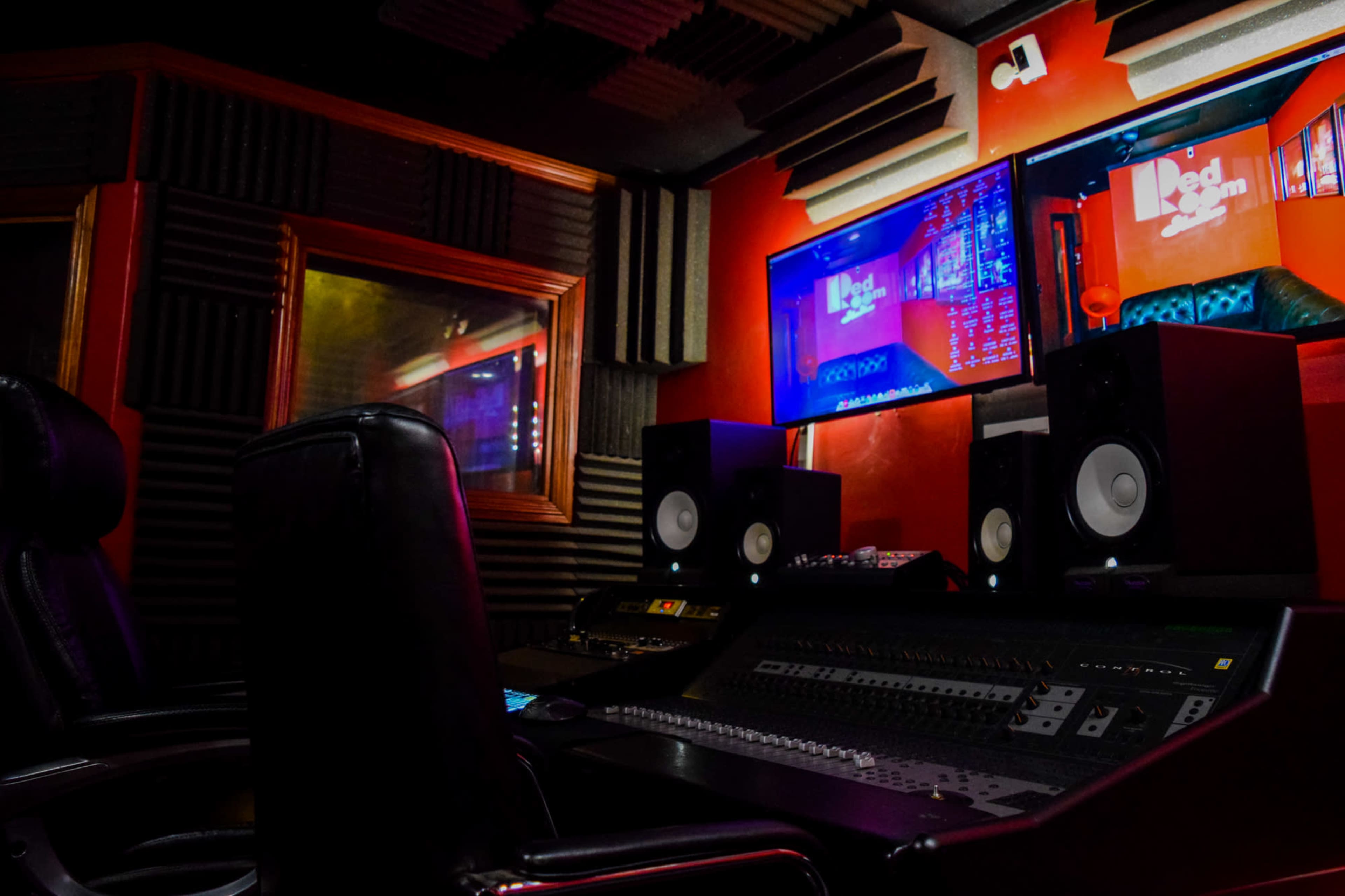 The image shows a music recording studio featuring a mixing console, two large speakers, and multiple screens displaying audio tracks.