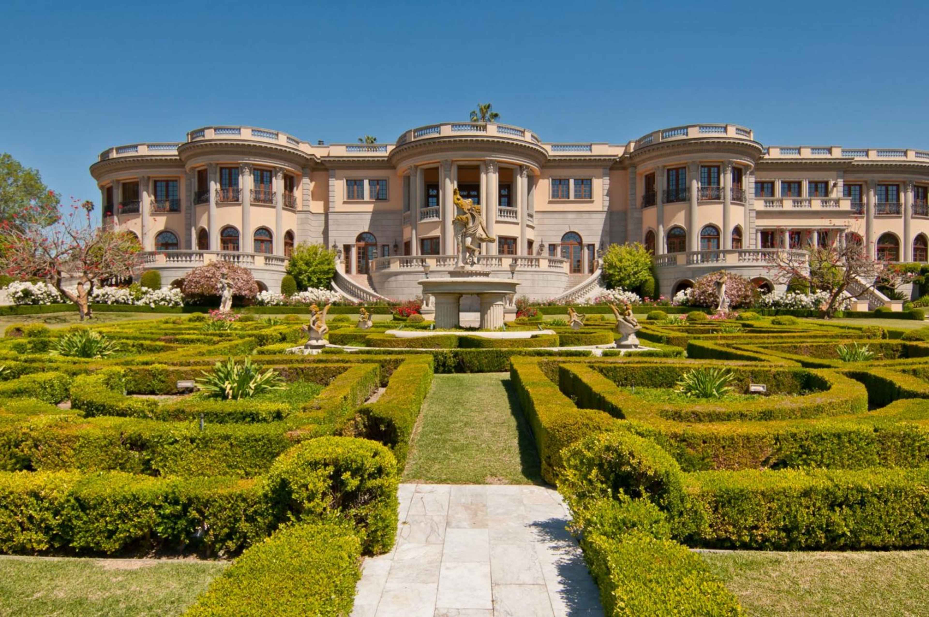 A large, elegant mansion is situated behind a meticulously designed garden featuring neatly trimmed hedges and decorative sculptures.