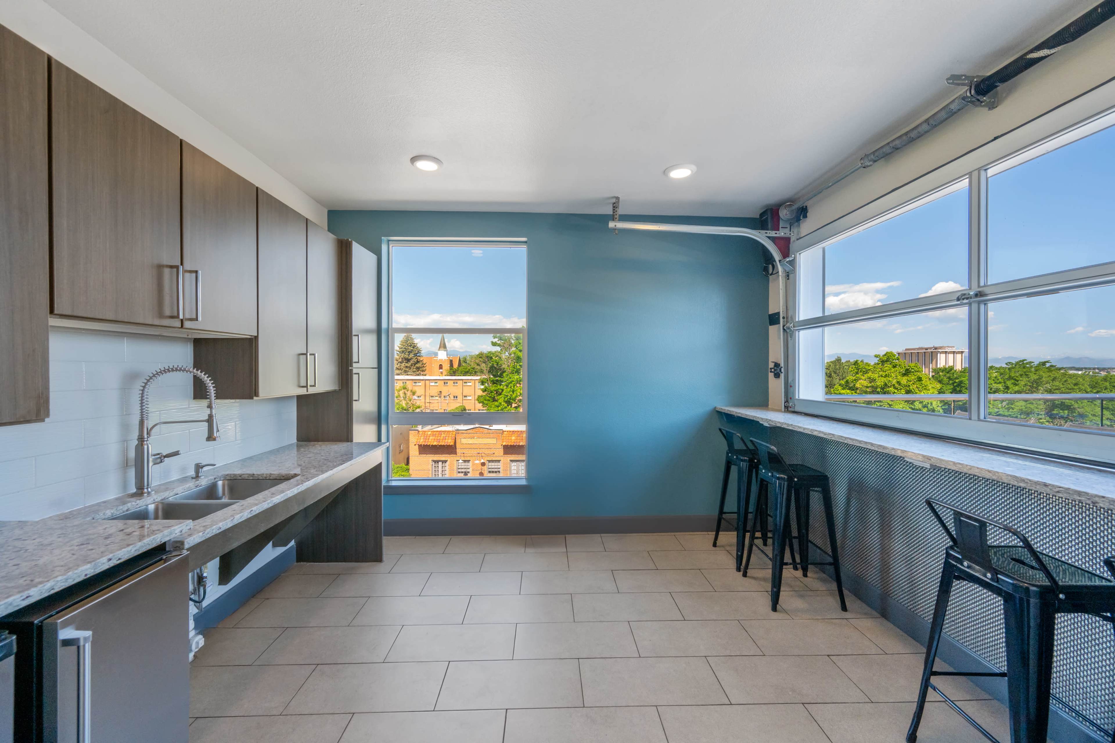 The image shows a modern kitchen with dark wooden cabinets, a sink, and a countertop, featuring large windows that provide a view of a building and trees outside.