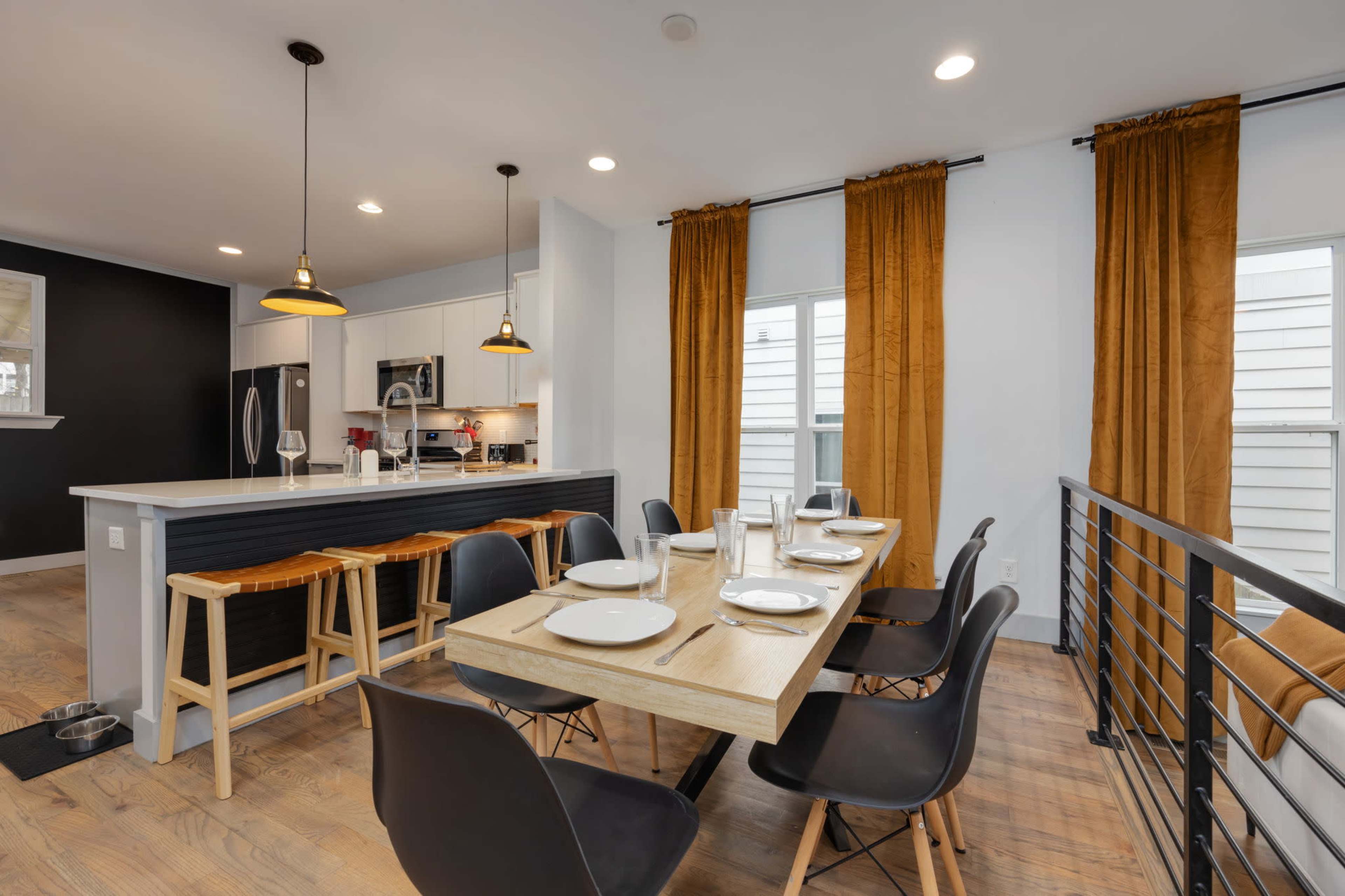 A modern dining area features a wooden table set for six, complemented by black chairs, and a kitchen with white cabinetry and a bar area with stools.