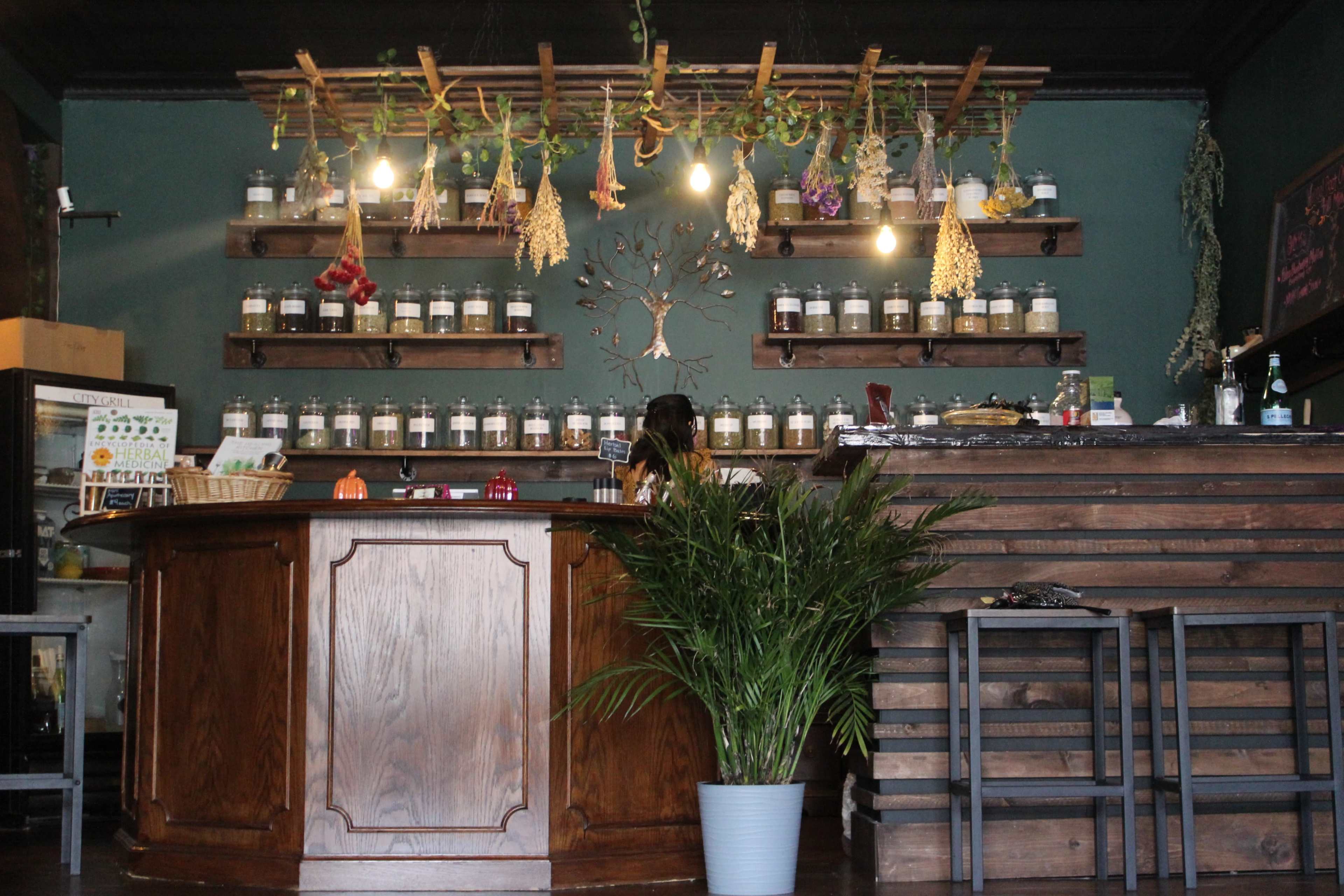 A wooden counter is positioned in front of a wall lined with jars and decorated with hanging herbs in a green-painted shop interior.