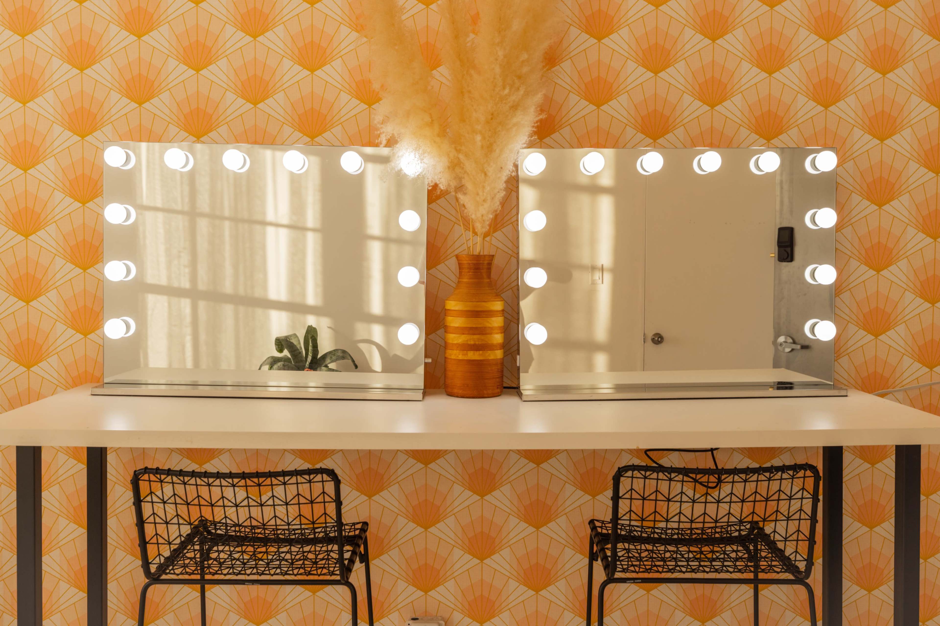 A table with two illuminated vanity mirrors and a vase of dried pampas grass against a patterned orange wallpaper.