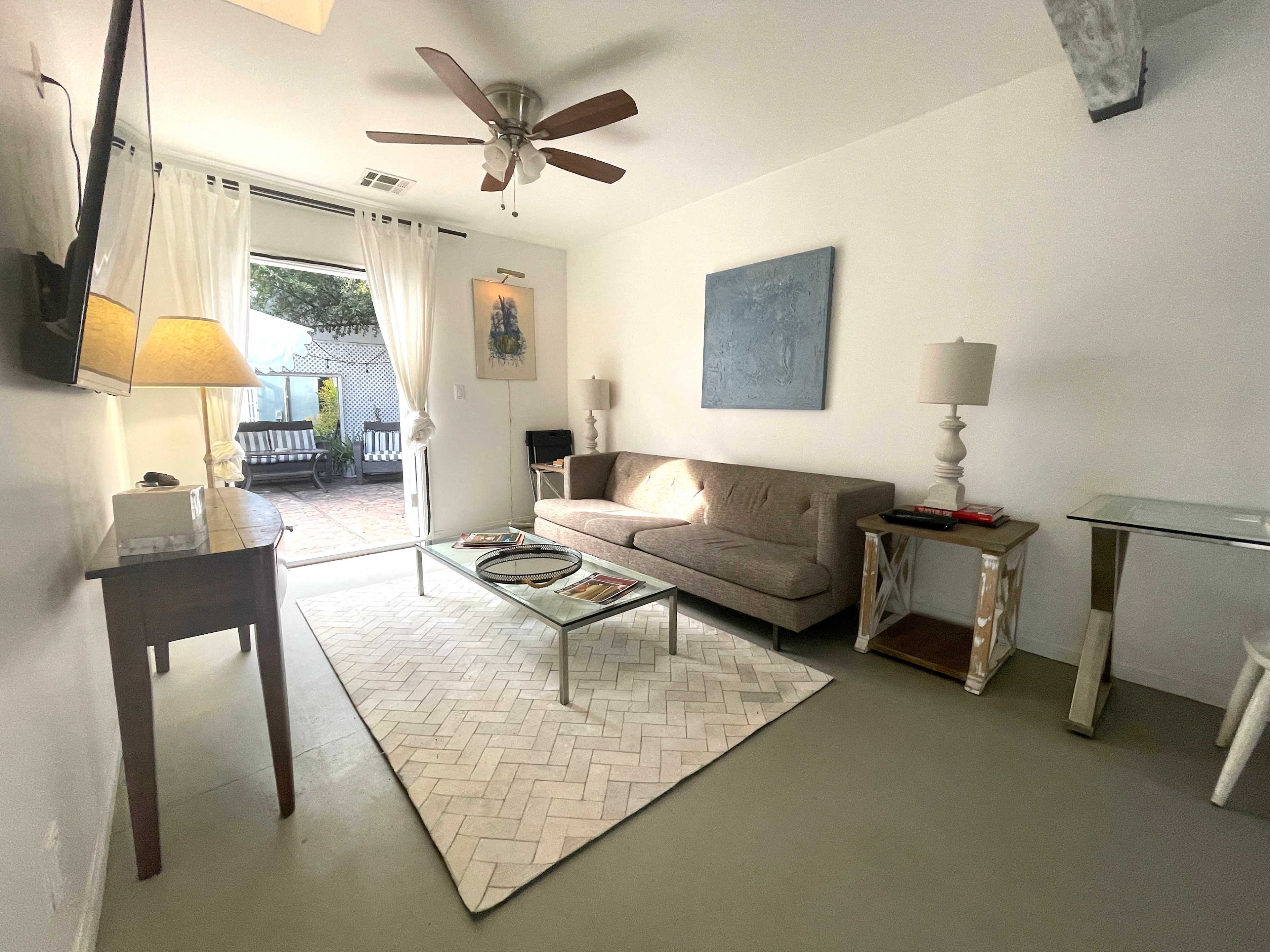 The image shows a bright living room with a sofa, a glass coffee table, two lamps, and a door leading to a patio area.