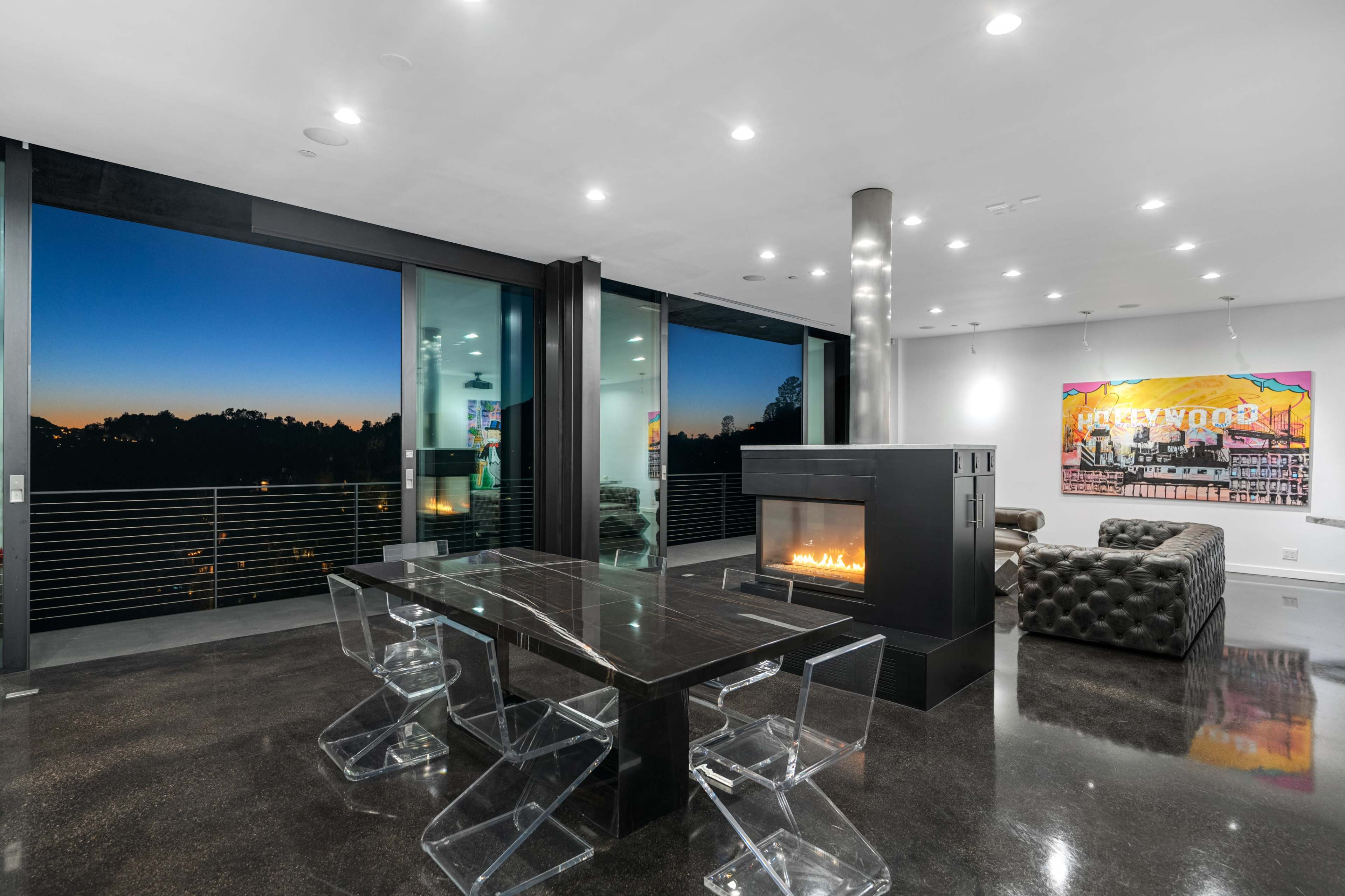 The image shows a modern living space with large windows overlooking a sunset, featuring a sleek dining table and a black fireplace.