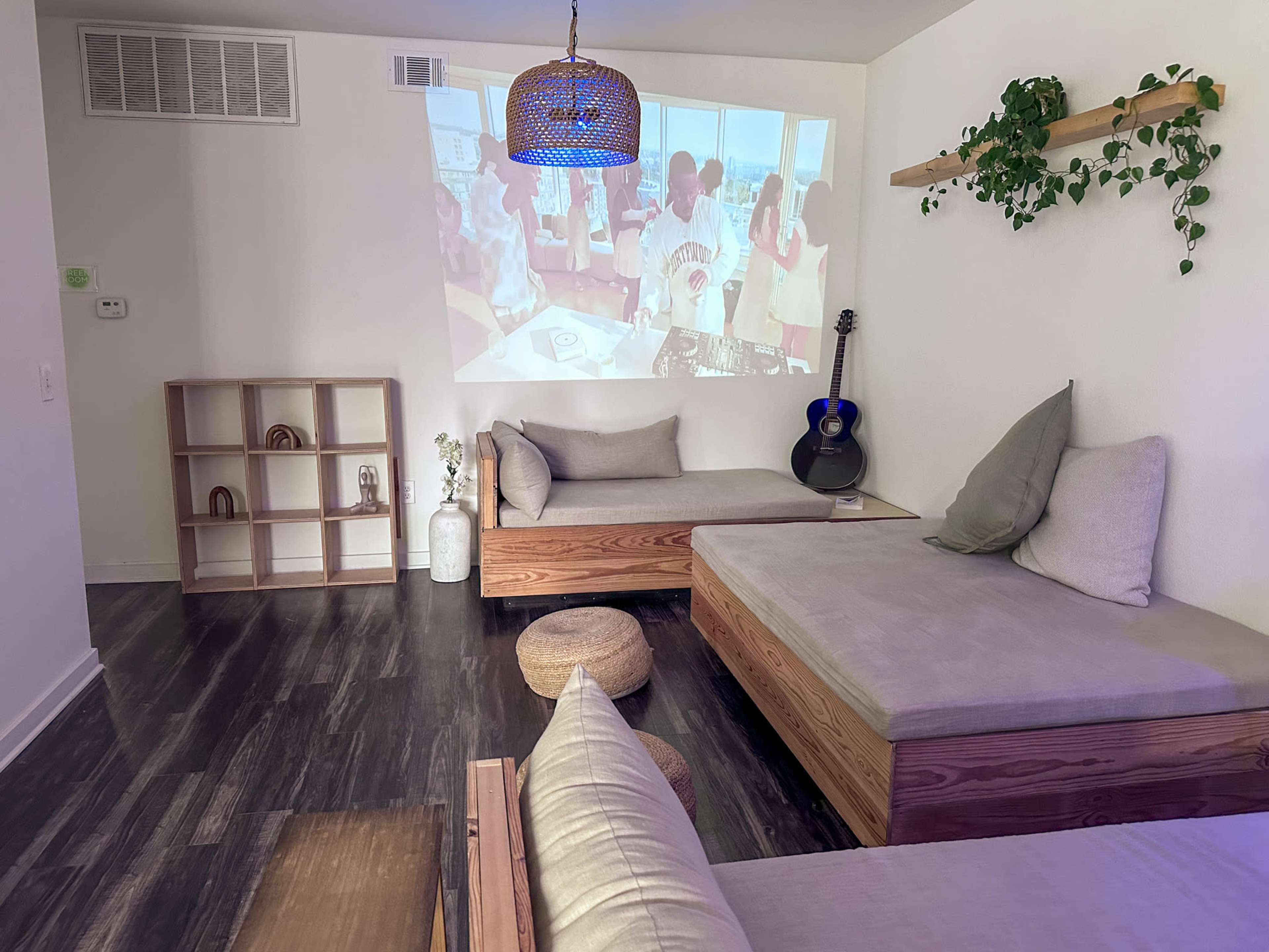 The image shows a modern, minimalist living space with two wooden couches, a guitar, and a projector displaying an indoor scene on the wall.