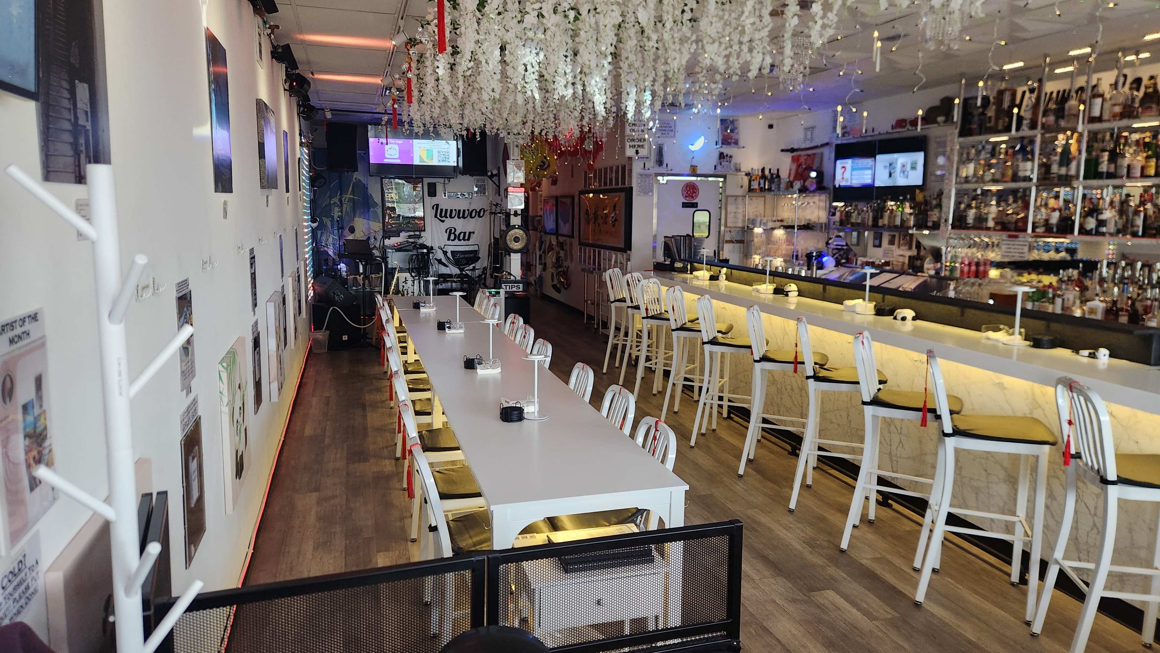 The image shows a modern bar and dining area with a long white table, white chairs, and a bar counter lined with various bottles.