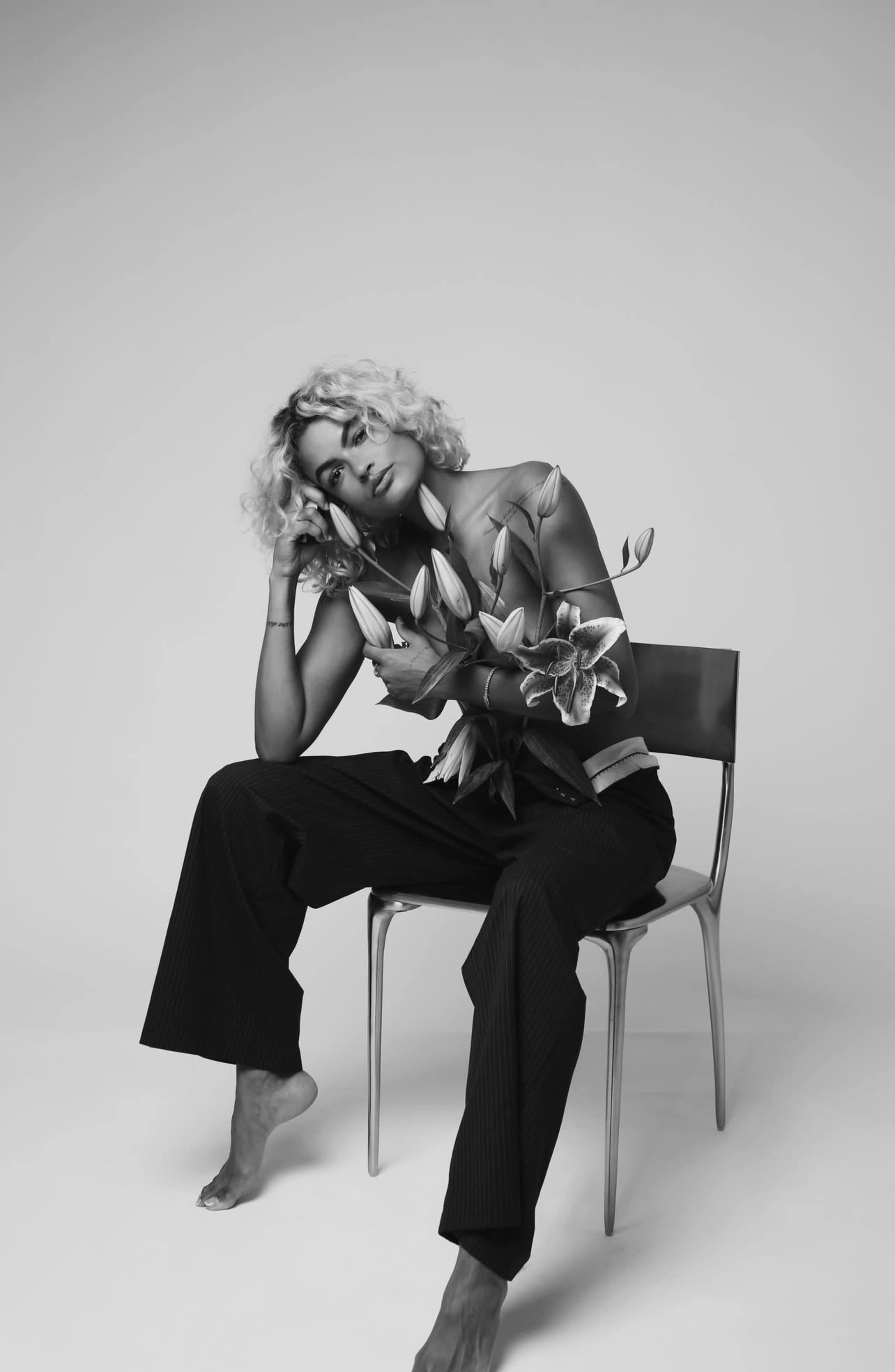 A person with curly hair sits on a chair, gently holding lilies against their arm.