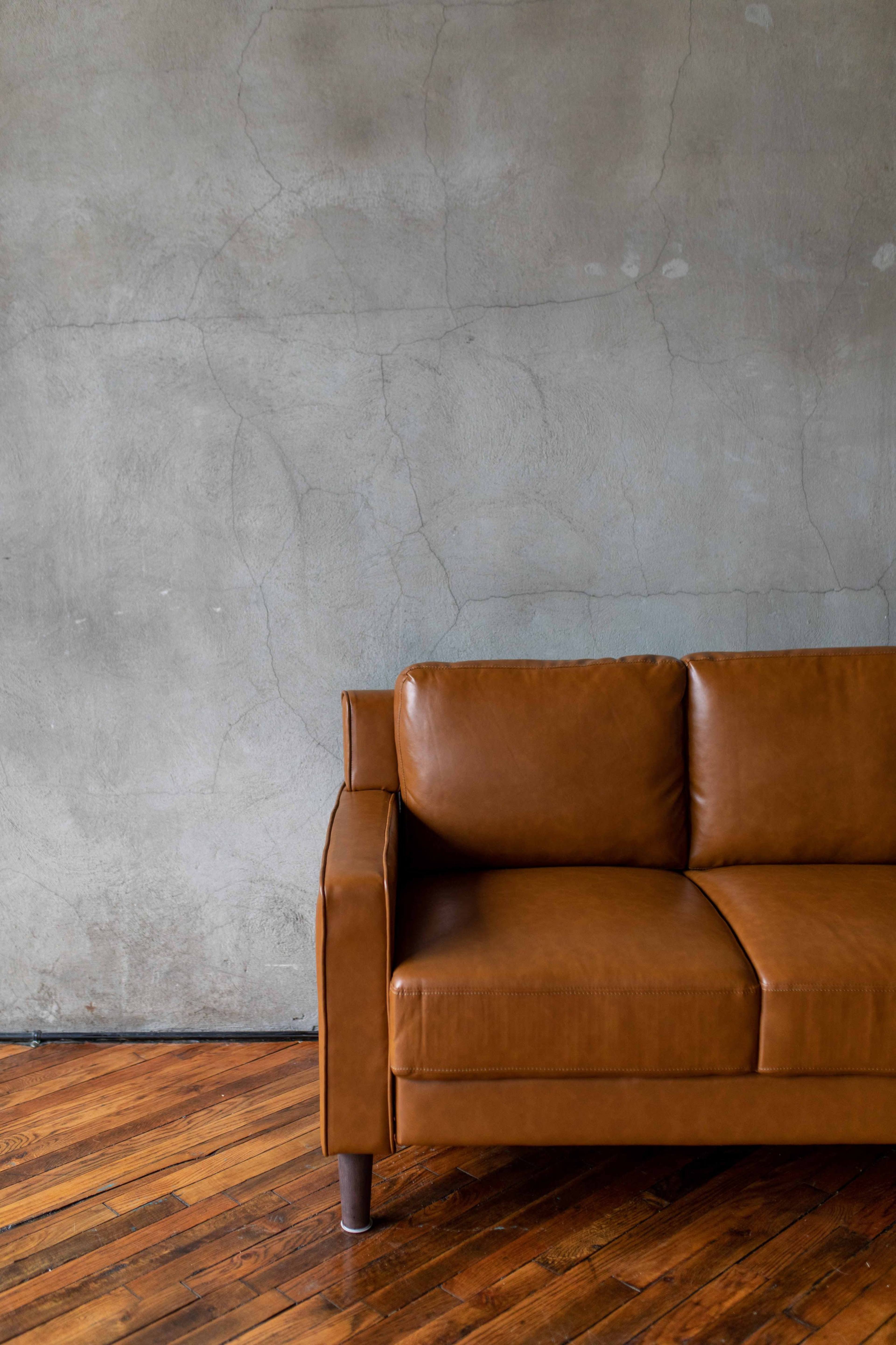 A brown leather sofa is positioned against a textured gray wall with visible cracks, on a wooden floor.