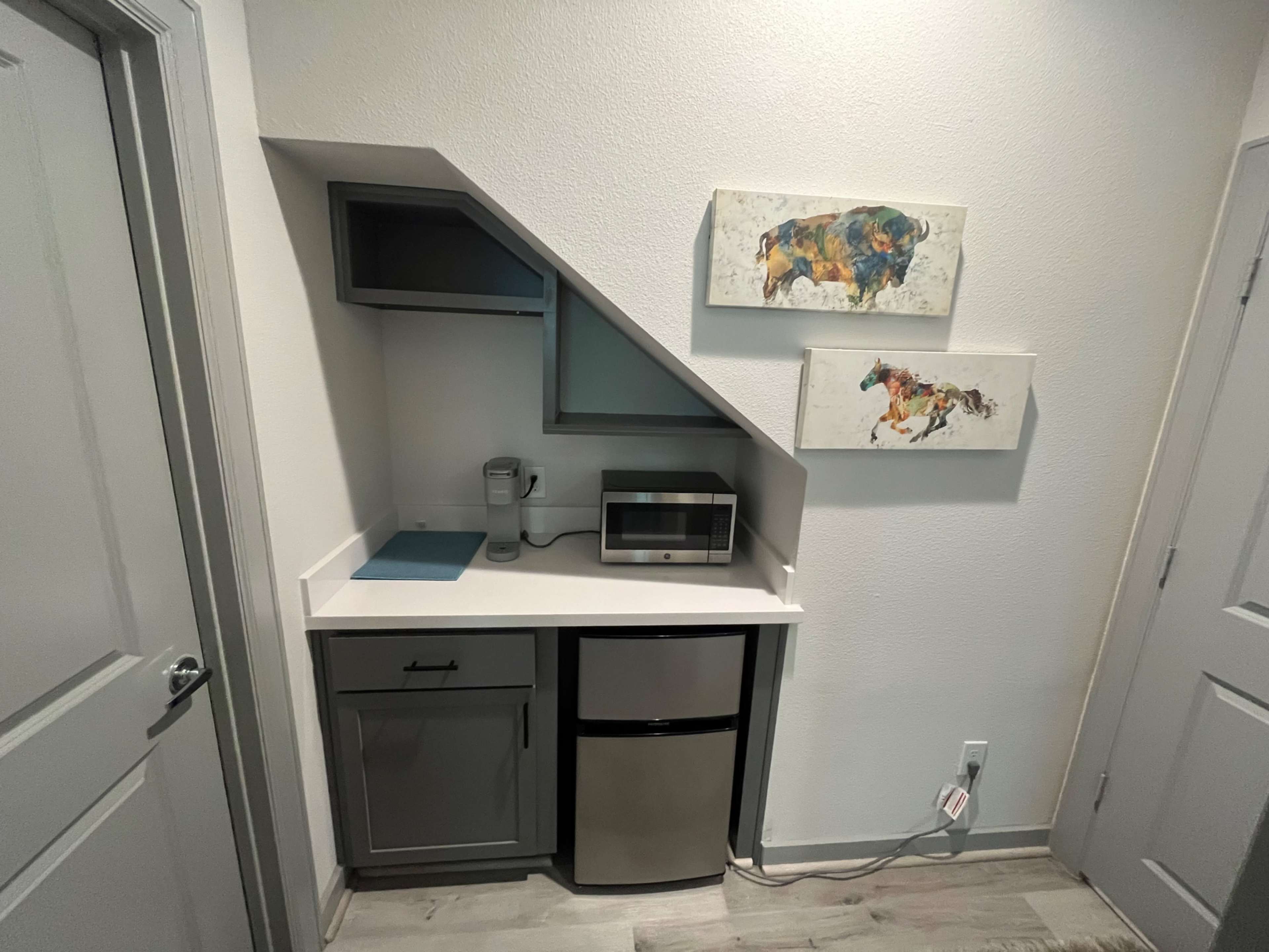 A compact kitchenette features a small refrigerator, microwave, and coffee maker, situated under an angled wall with two framed artworks above.