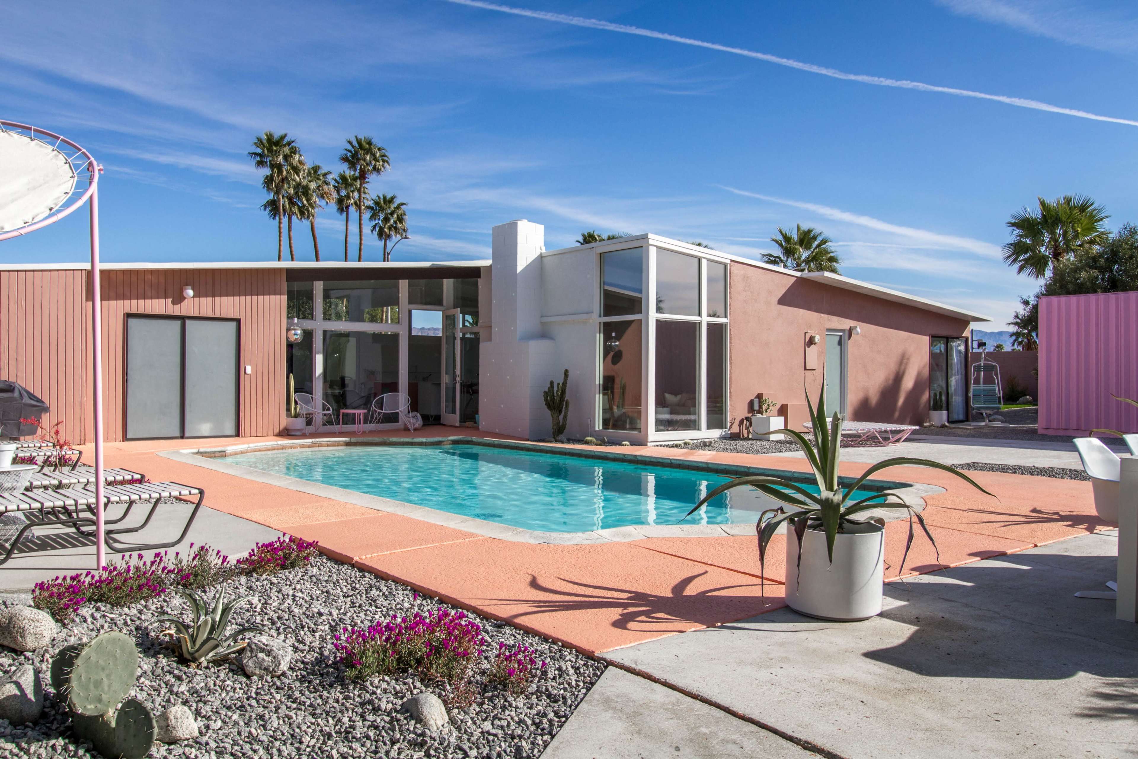 A modern house with large windows surrounds a swimming pool, featuring a pink exterior and desert landscaping with palm trees.