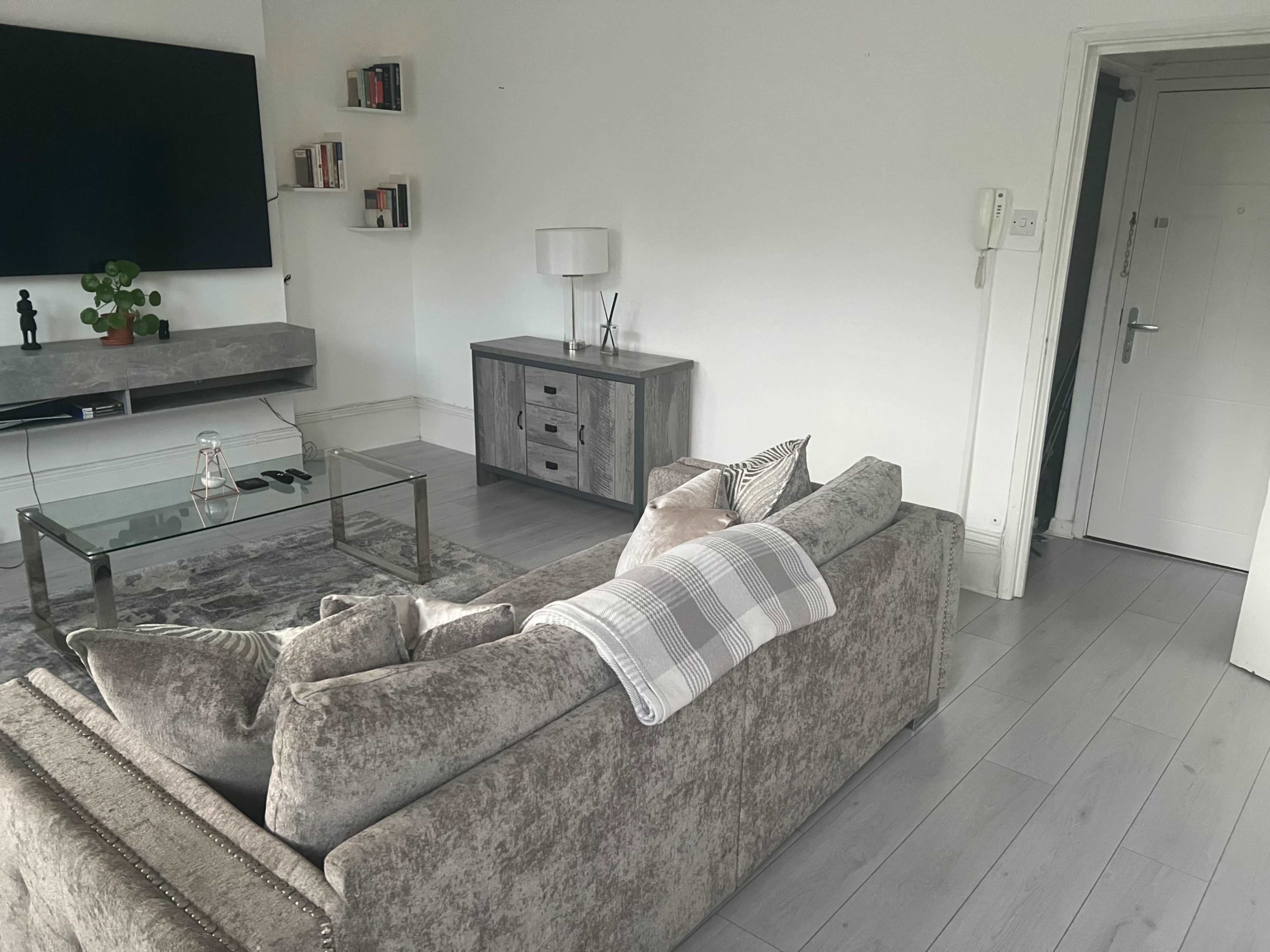 A living room features a gray sofa, a glass coffee table, a television on the wall, and a console table near the entrance.