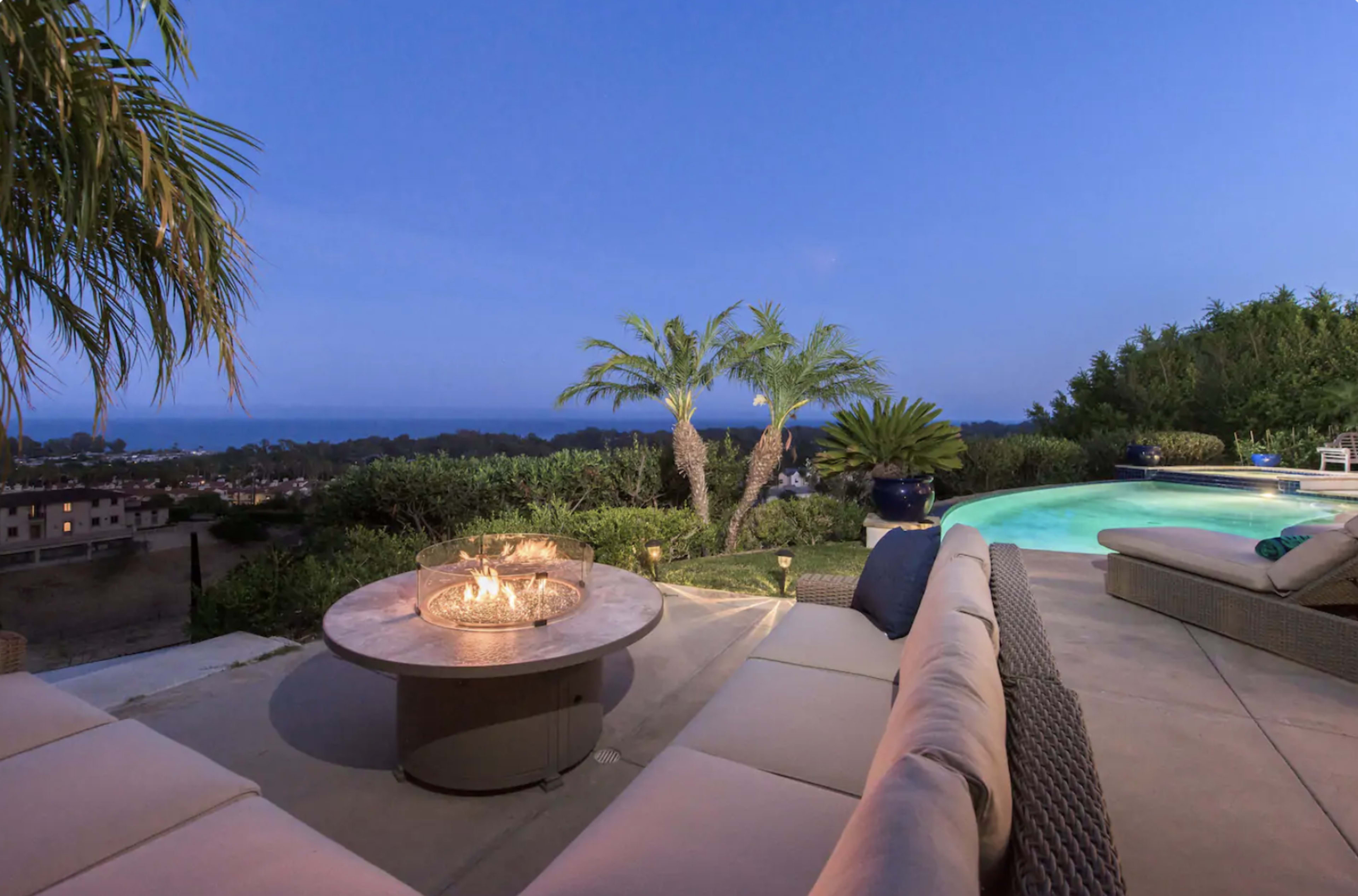 A patio area with a circular fire pit and seating, overlooking a swimming pool and a view of the ocean at dusk.