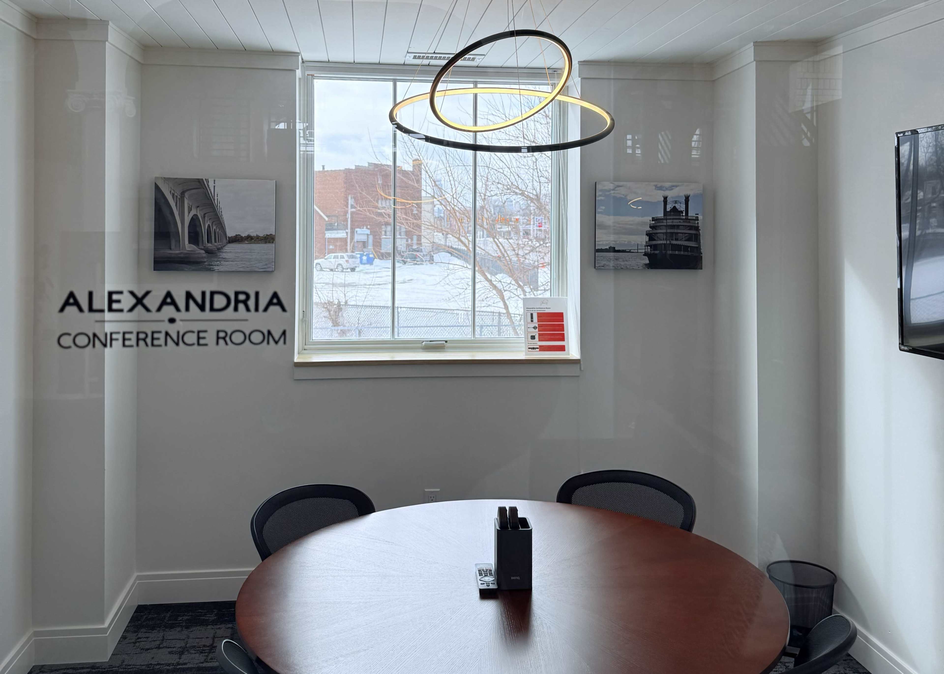 The image features a conference room with a round wooden table, four black chairs, and a large window displaying a view of an outdoor area.