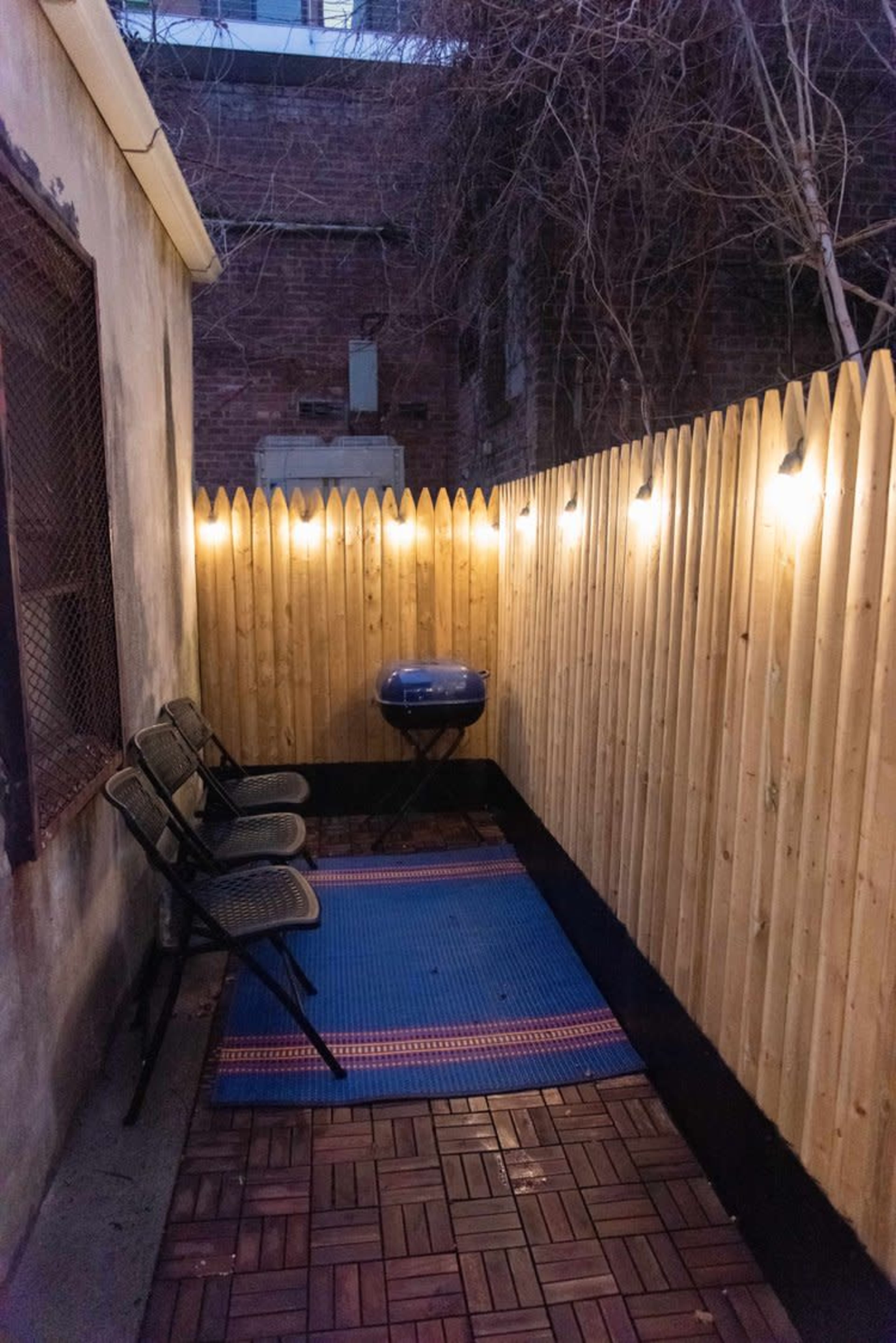 A narrow outdoor space with a wooden fence, two black chairs, a blue grill, and string lights overhead.
