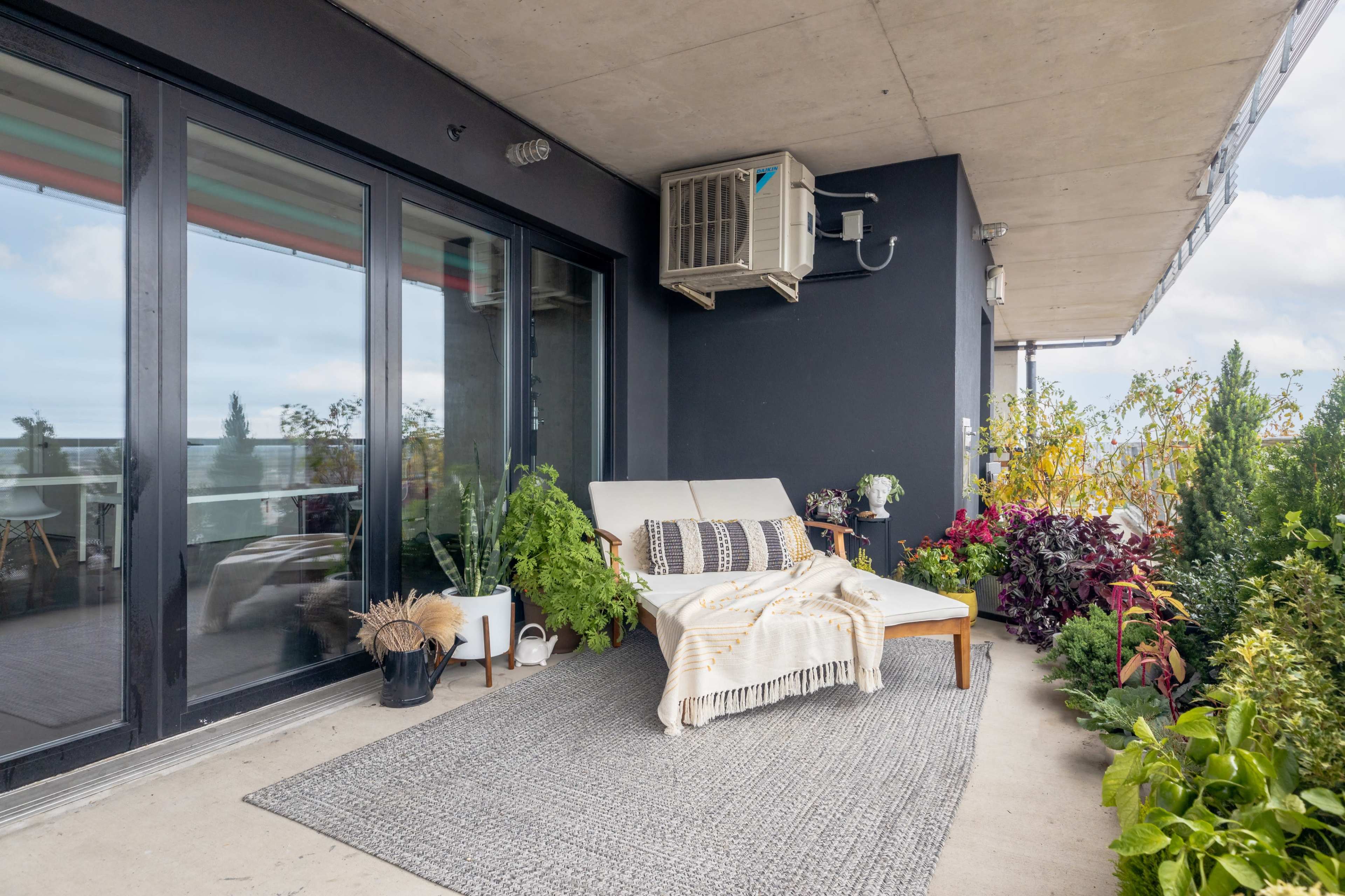 The image shows a cozy balcony with a lounge chair, a textured rug, various potted plants, and an air conditioning unit on the wall.
