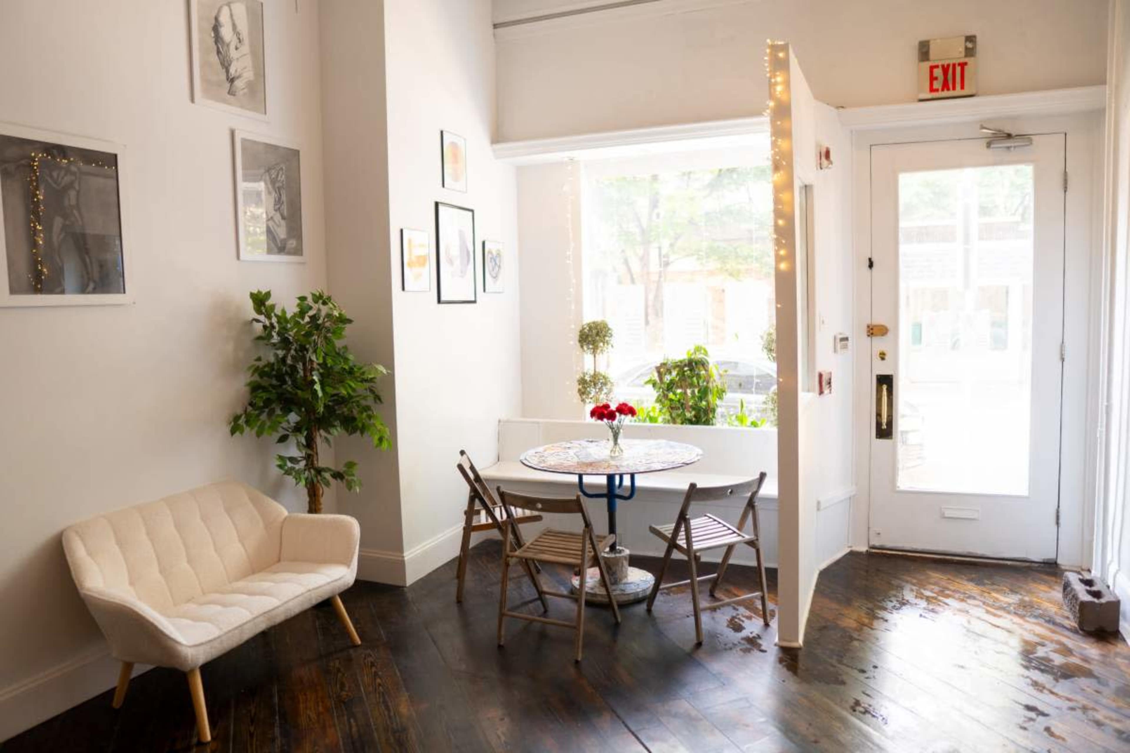 A small, bright room featuring a seating area with a light-colored chair and a round table adorned with a vase of red flowers, beside a window.