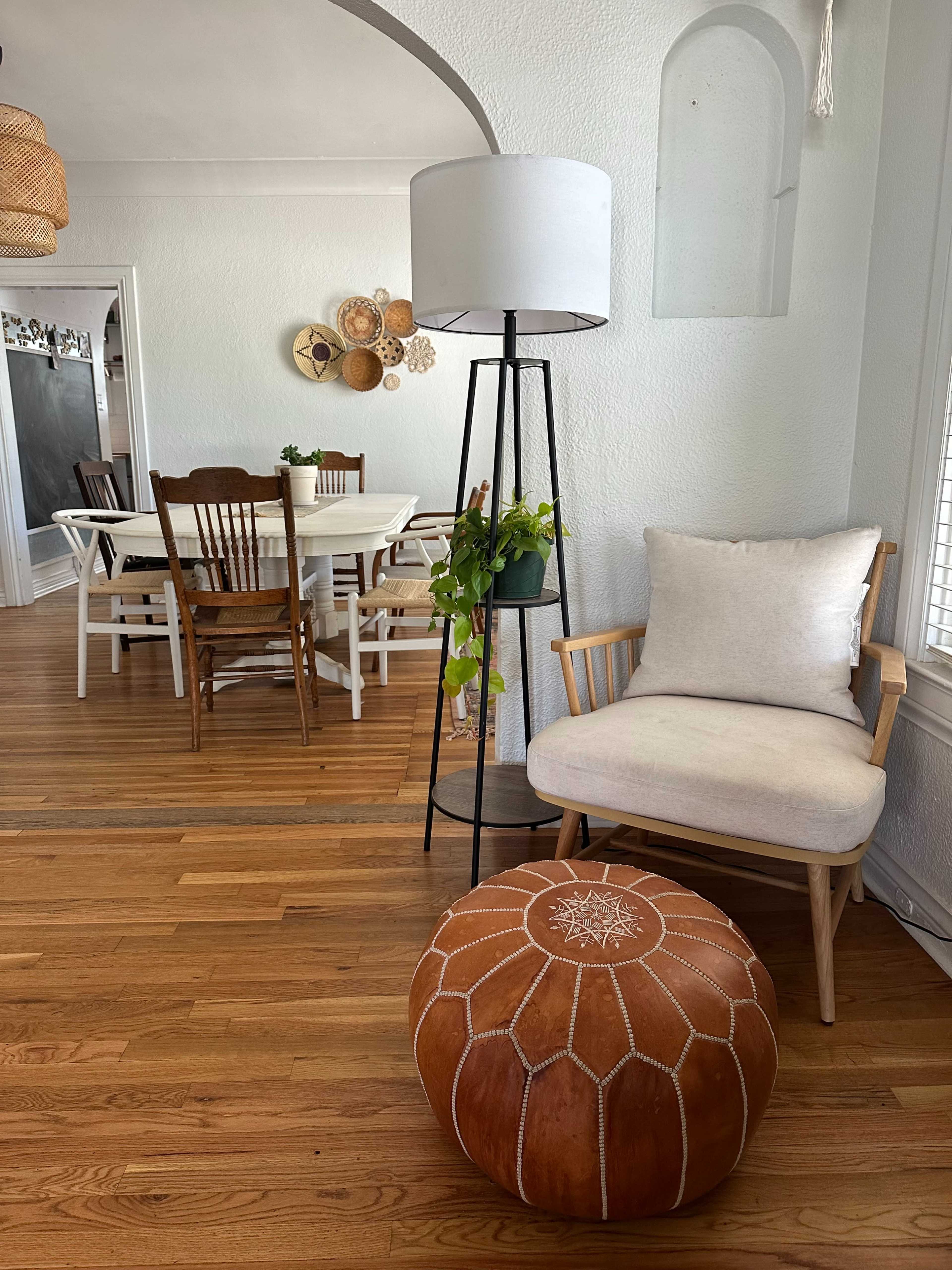 The image shows a cozy living area featuring a chair with a cushion, a patterned pouf, a standing lamp, and a dining table in the background.