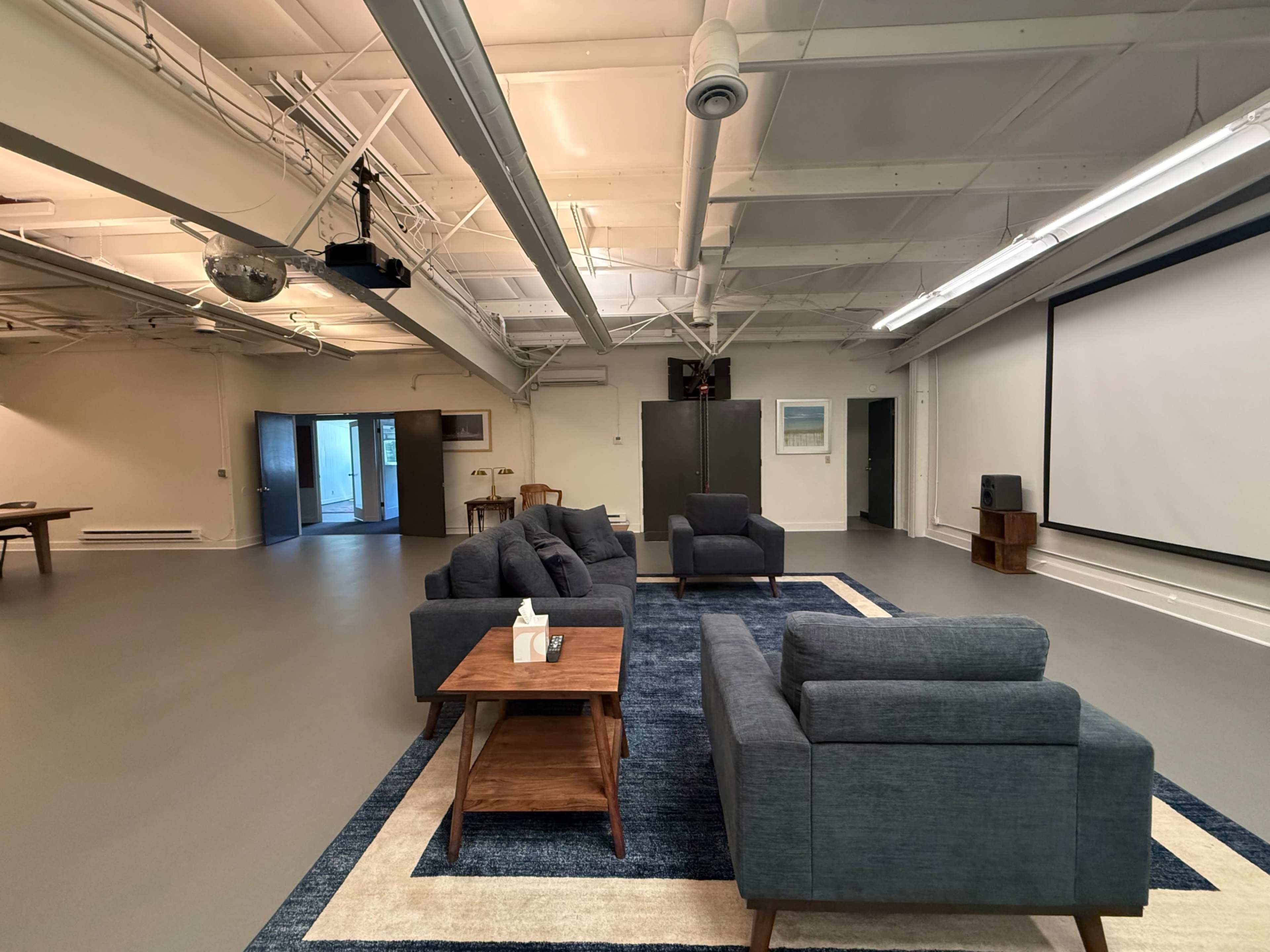 The image shows a spacious room with gray walls and a large central area furnished with a seating arrangement of blue sofas and a wooden coffee table atop a patterned rug, alongside a wall-mounted screen.
