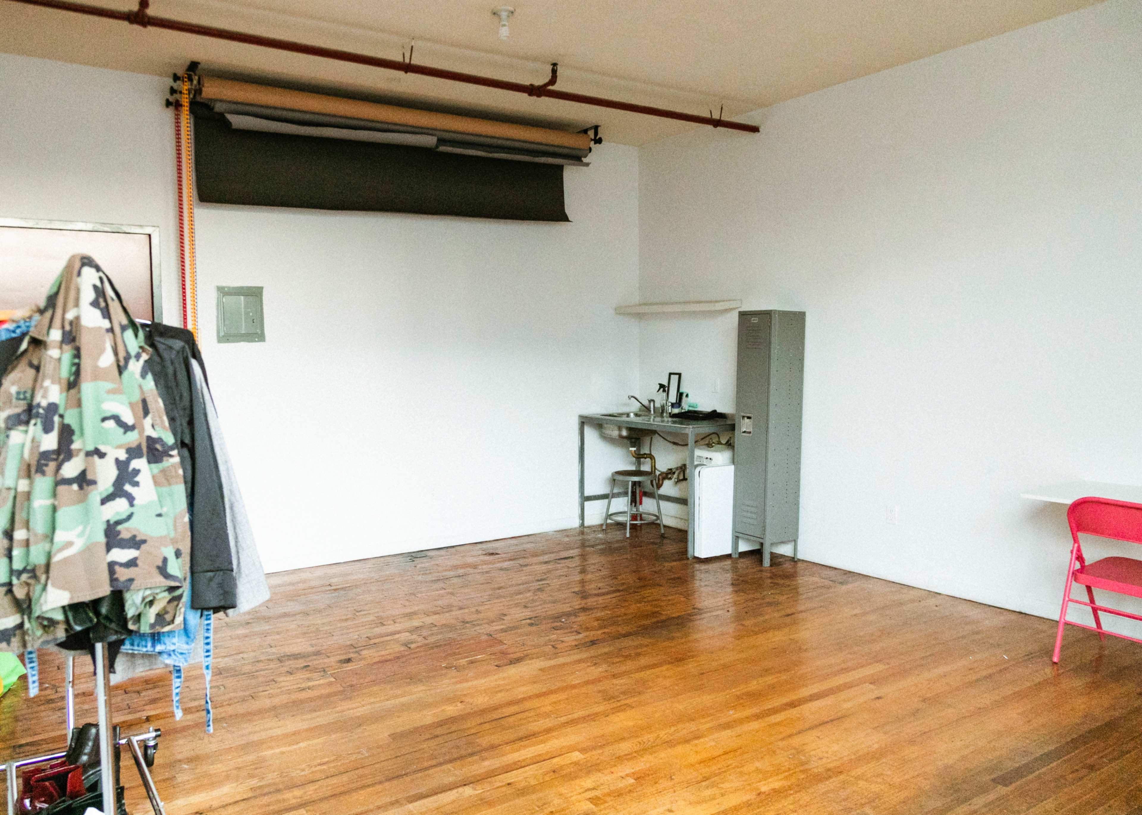 An empty room with wooden floors features a gray metal filing cabinet, a desk with equipment, and a clothing rack holding camouflage garments.