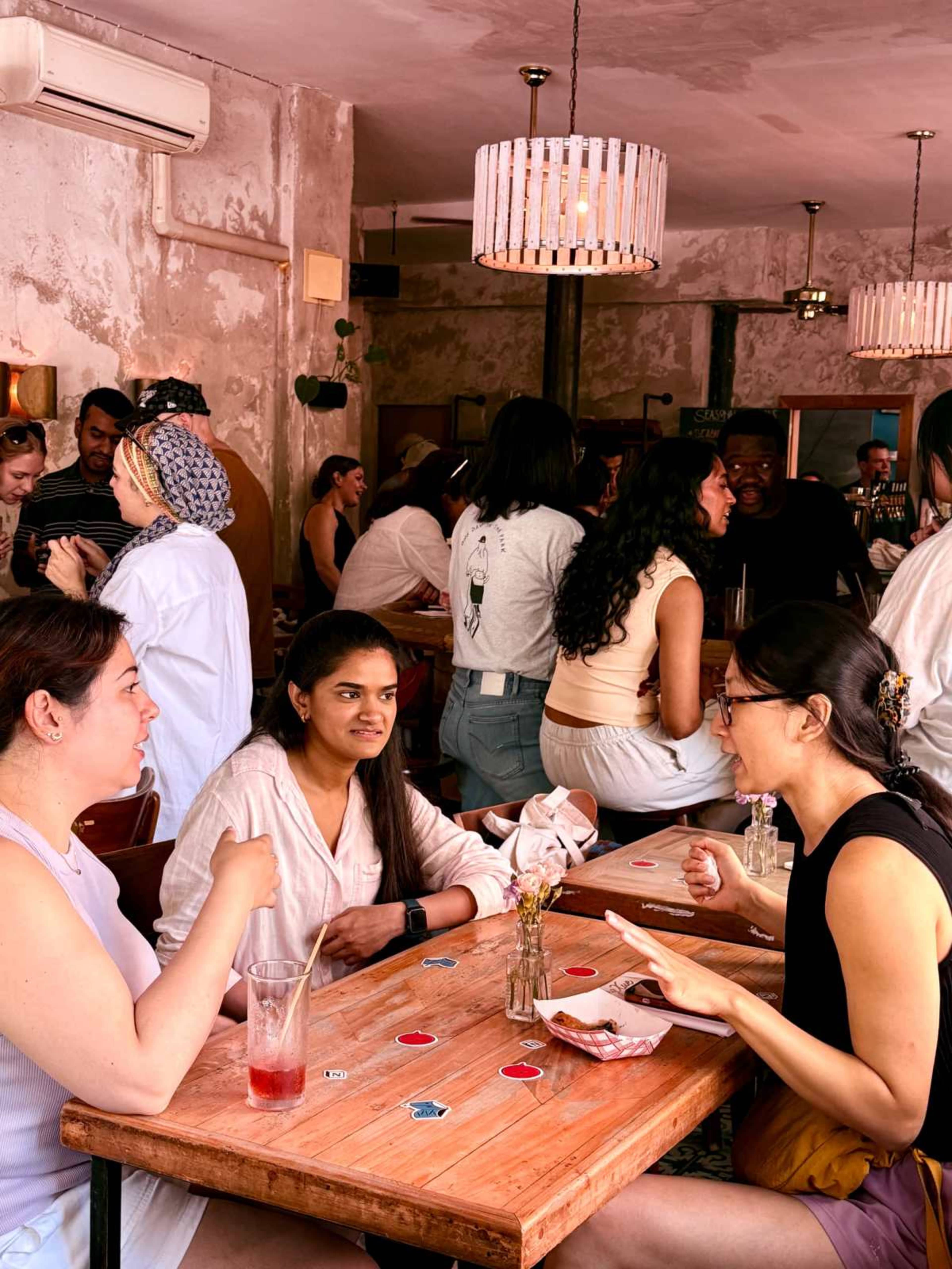 A busy café interior features several groups of people seated at wooden tables, chatting and enjoying drinks in a brightly lit space with decorative lighting.