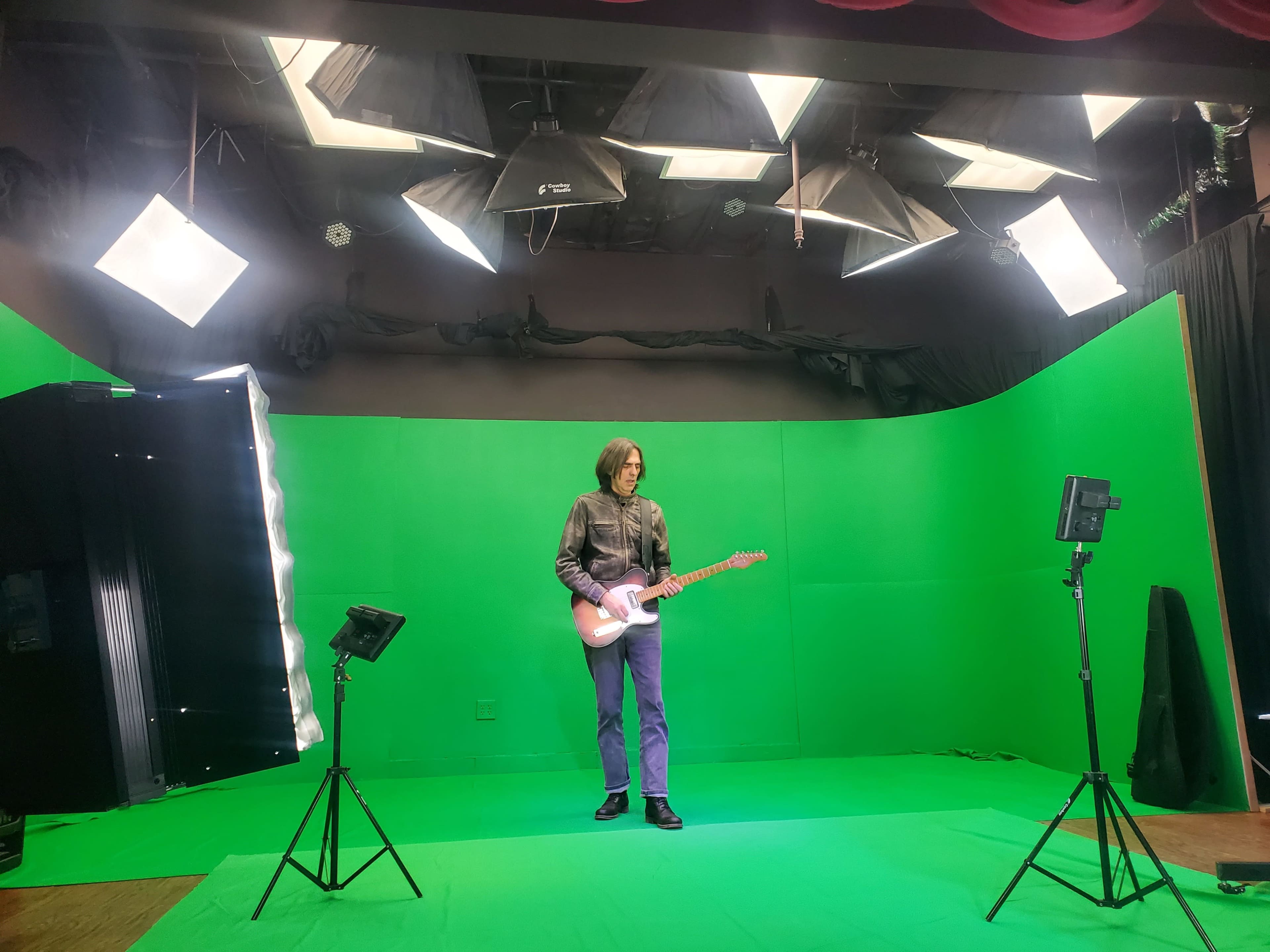 A person stands on a green backdrop set, holding an electric guitar, surrounded by studio lighting equipment.