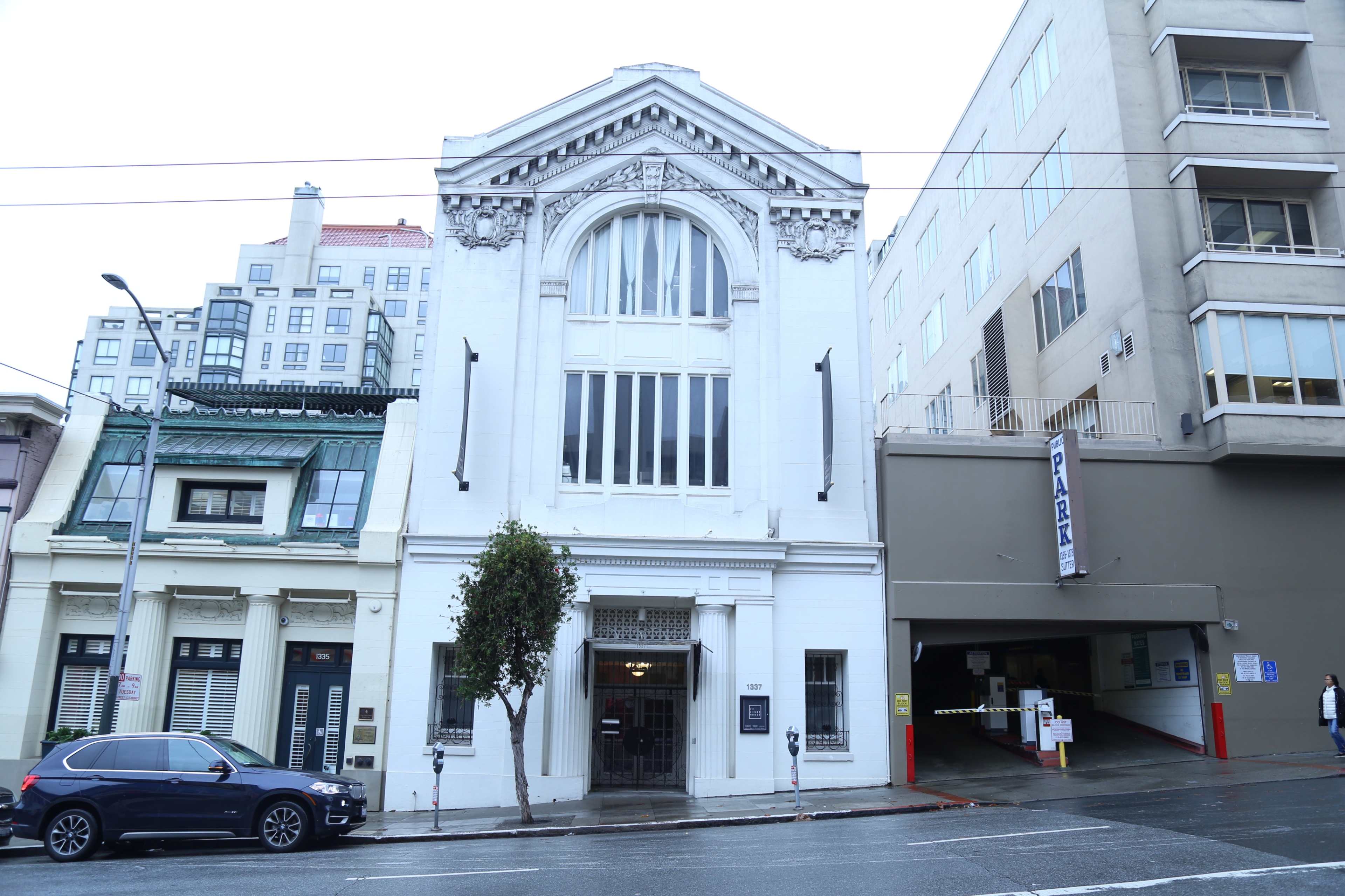 A white, historic building with large front windows, located next to a modern parking garage in an urban setting.