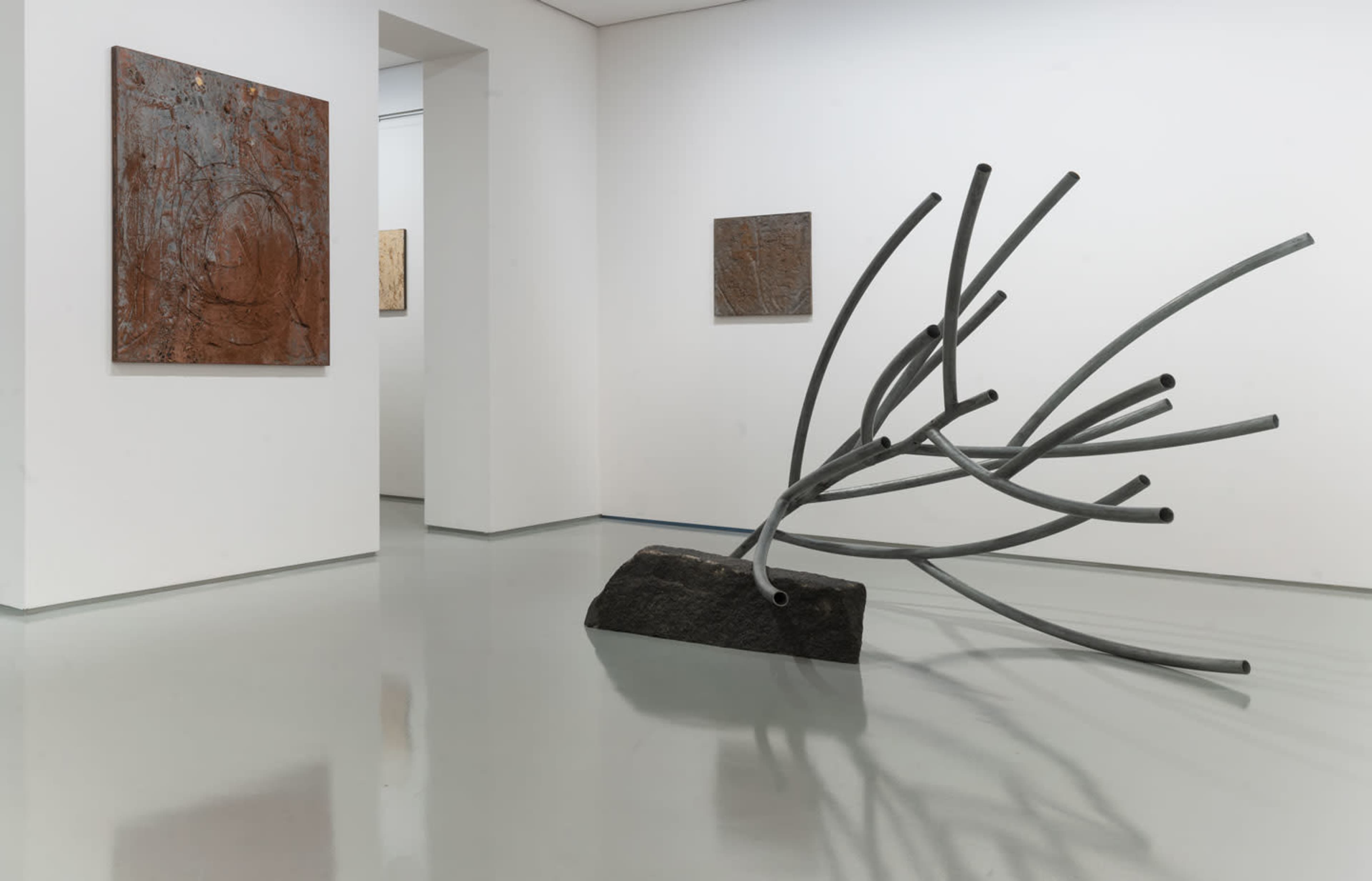 The image shows an art gallery with abstract paintings on the walls and a large sculpture made of gray pipes leaning against a black rectangular base in the foreground.