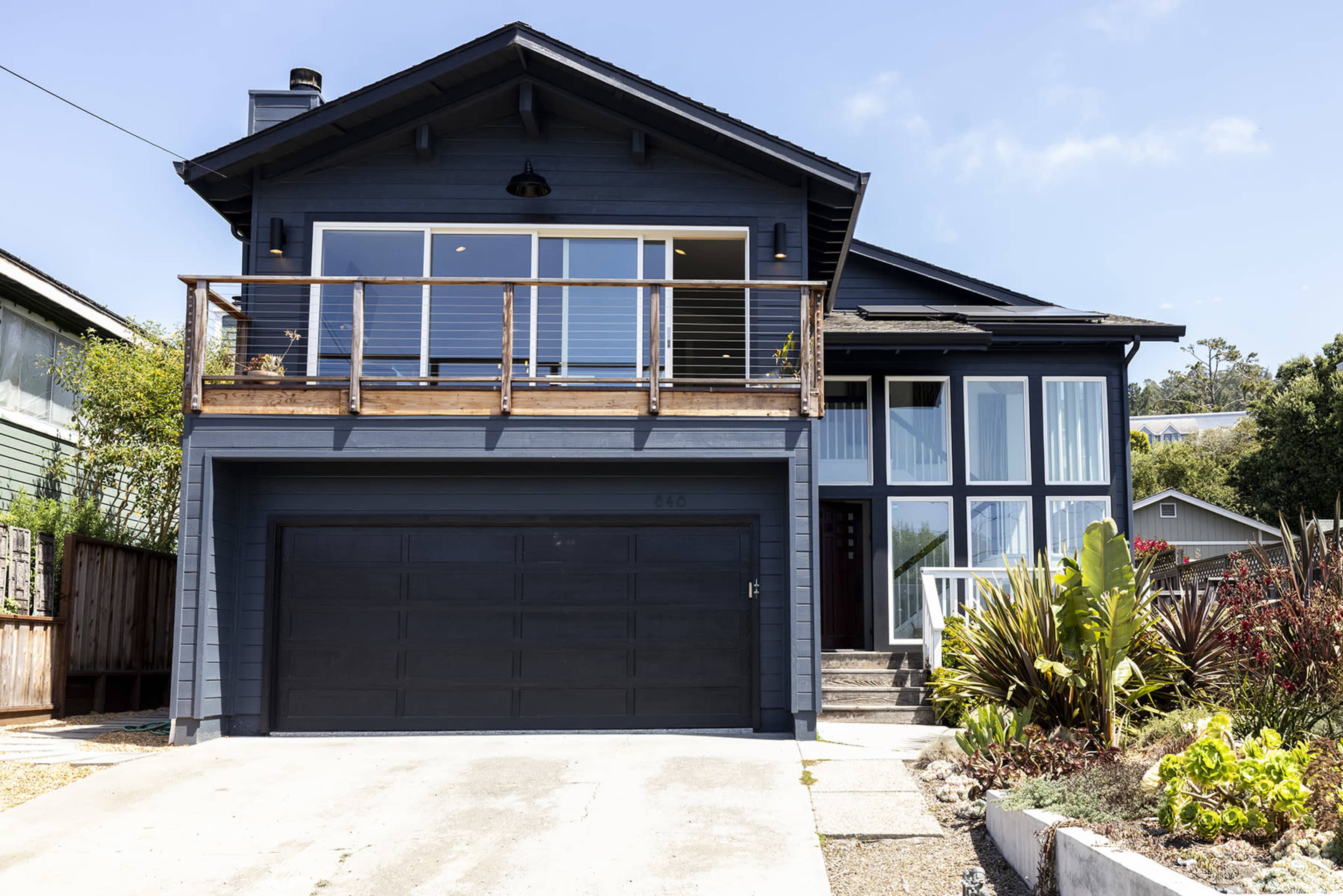A modern, two-story blue house with a balcony over the garage, surrounded by greenery.