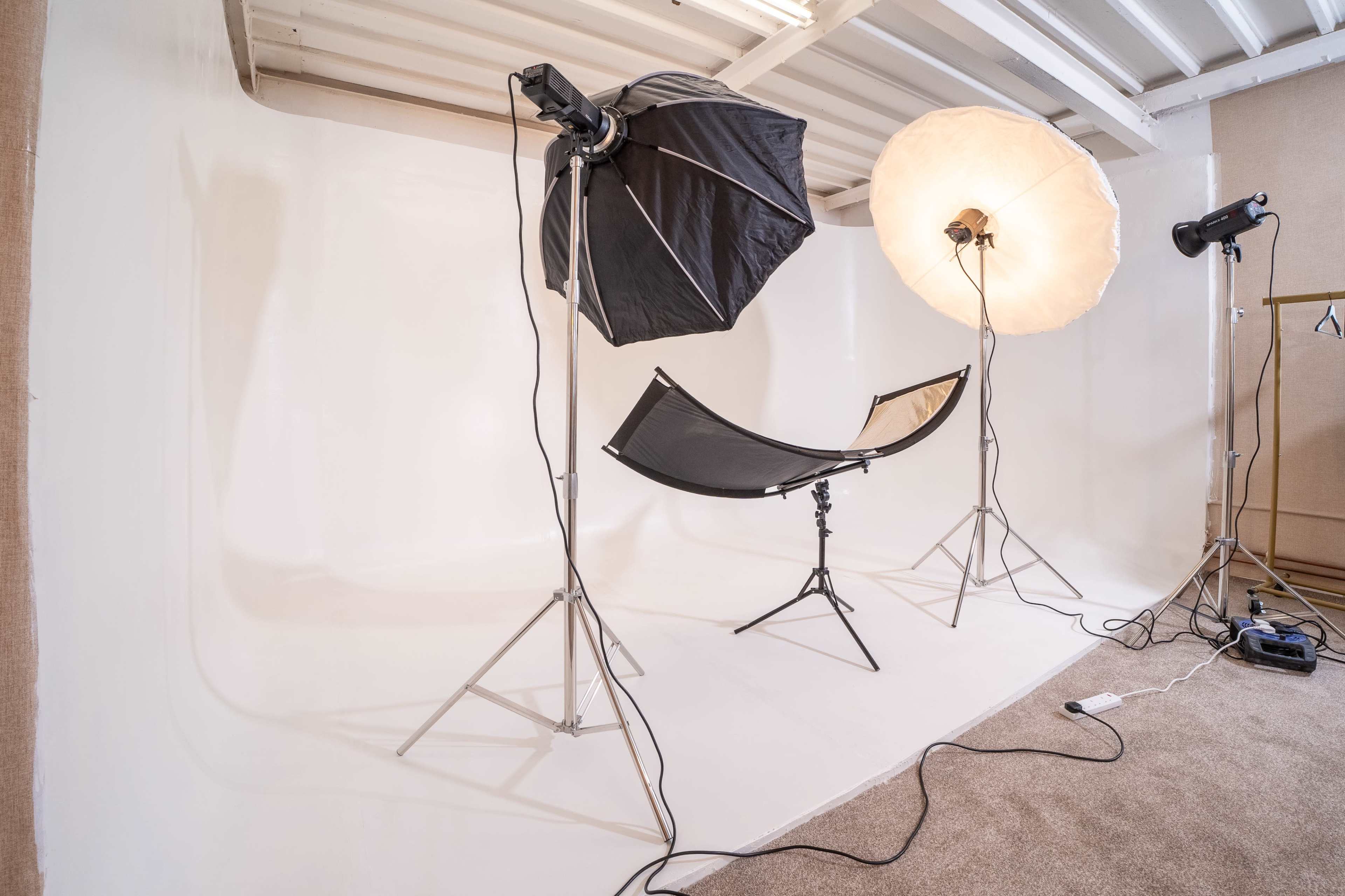 The image shows a photography studio setup with two light sources, a black umbrella softbox, a large round softbox, and a black folding chair positioned on a seamless white backdrop.