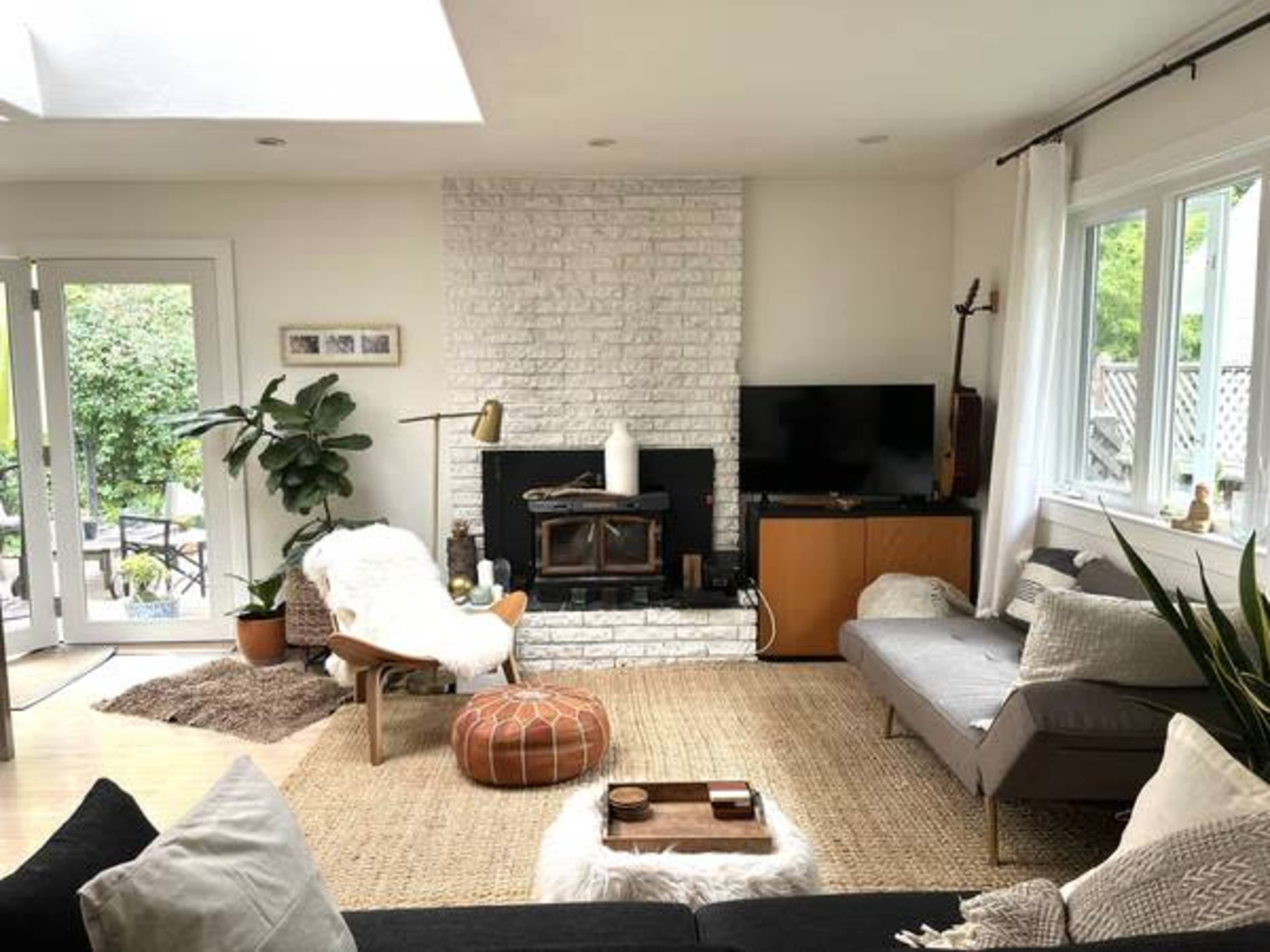 The living room features a white-brick fireplace, a television, and a mix of seating arrangements with natural elements and a large woven area rug.