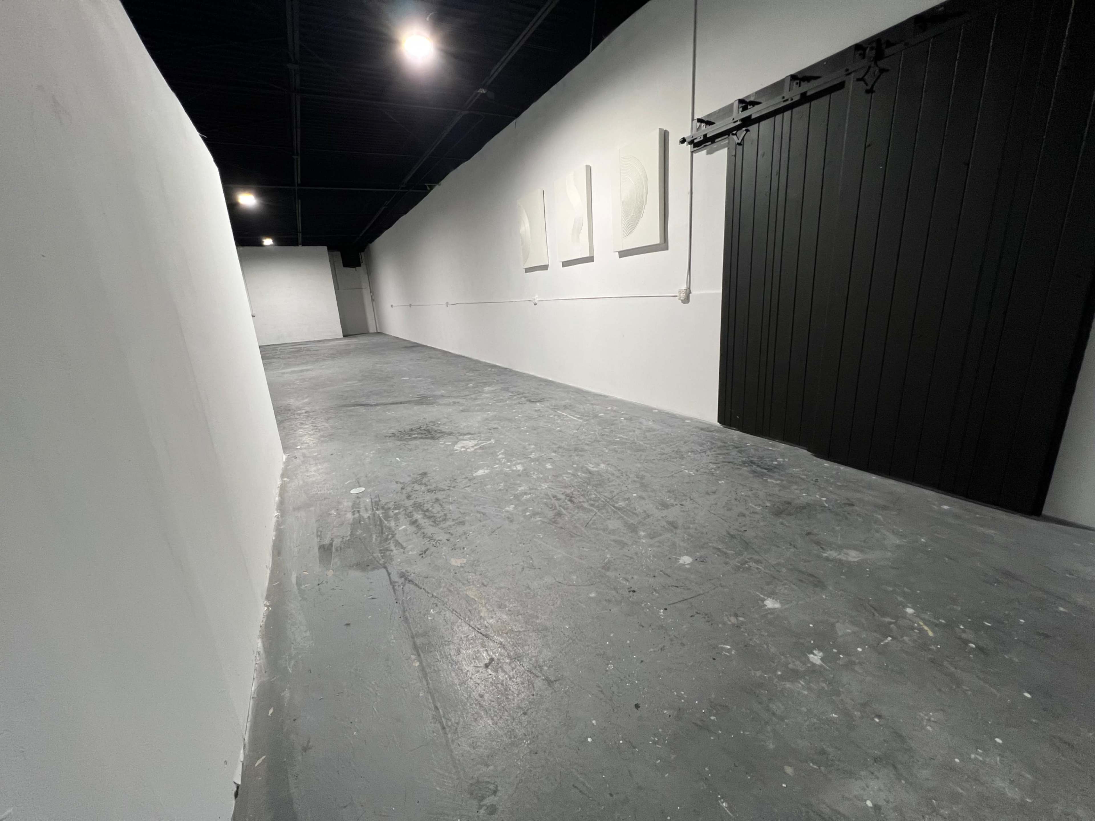 The image shows a long, narrow hallway with a gray concrete floor and white walls, featuring a black barn door at one end and several framed artworks hanging on the wall.