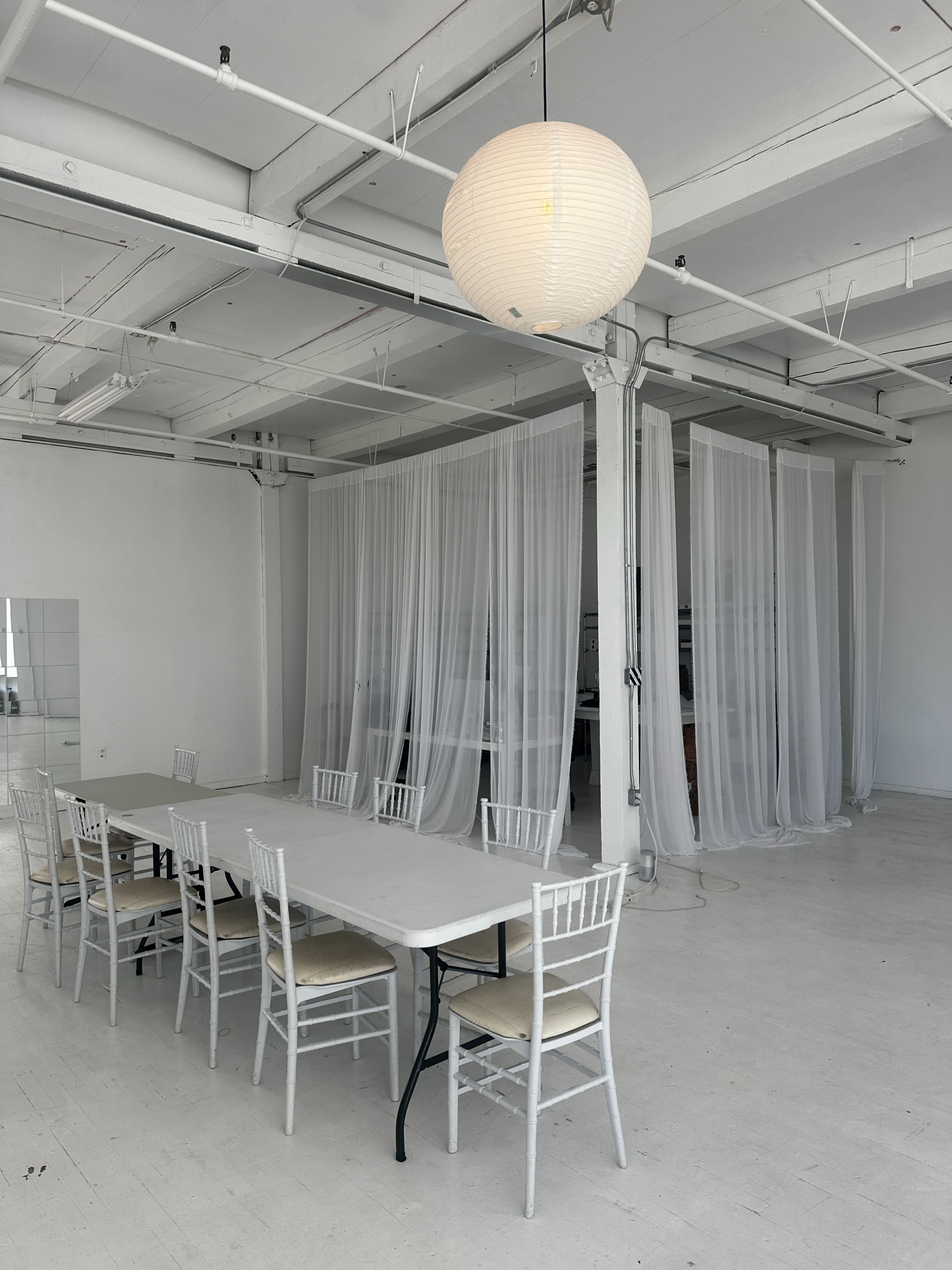 A long, white table with several chairs is set in a spacious room featuring sheer curtains and bright overhead lighting.