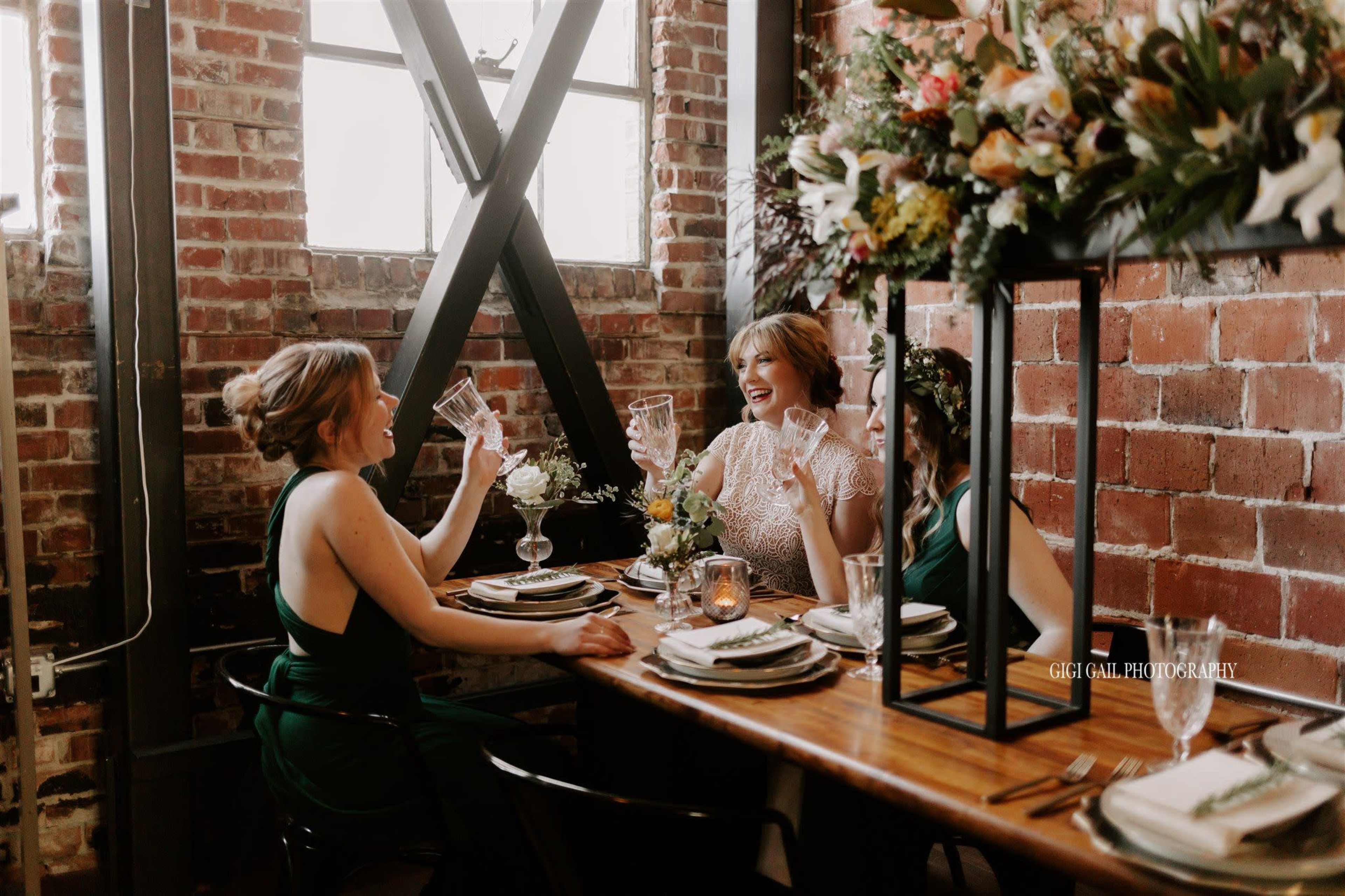 Three women toast with glasses at a wooden table set with plates and flowers inside a rustic brick-walled venue.