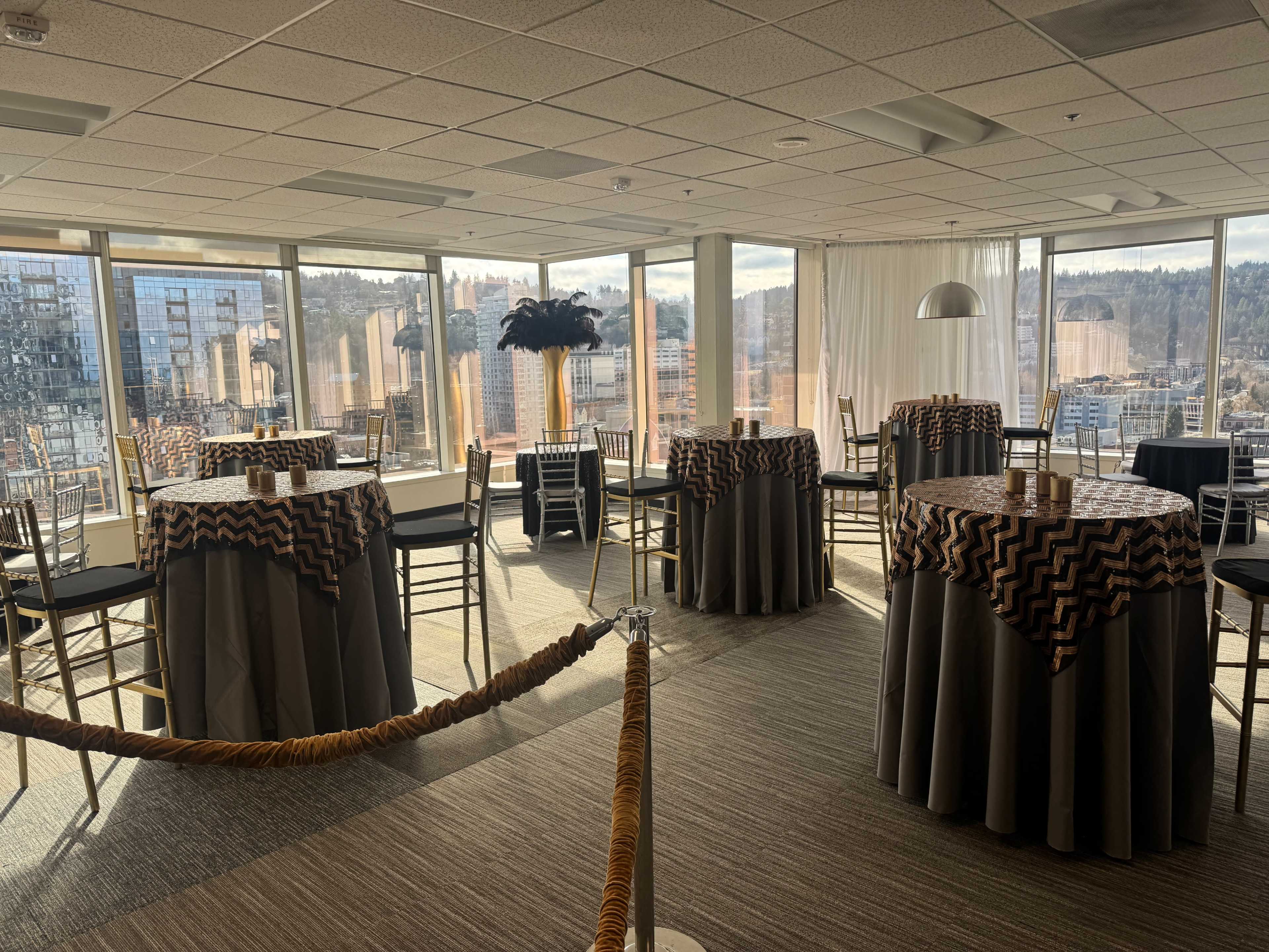 The image shows a well-lit event space with several round tables covered in patterned tablecloths and a view of a city skyline through large windows.